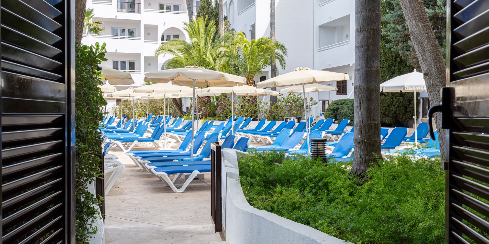 Resort pool area with blue sun loungers, beige umbrellas, and palm trees.