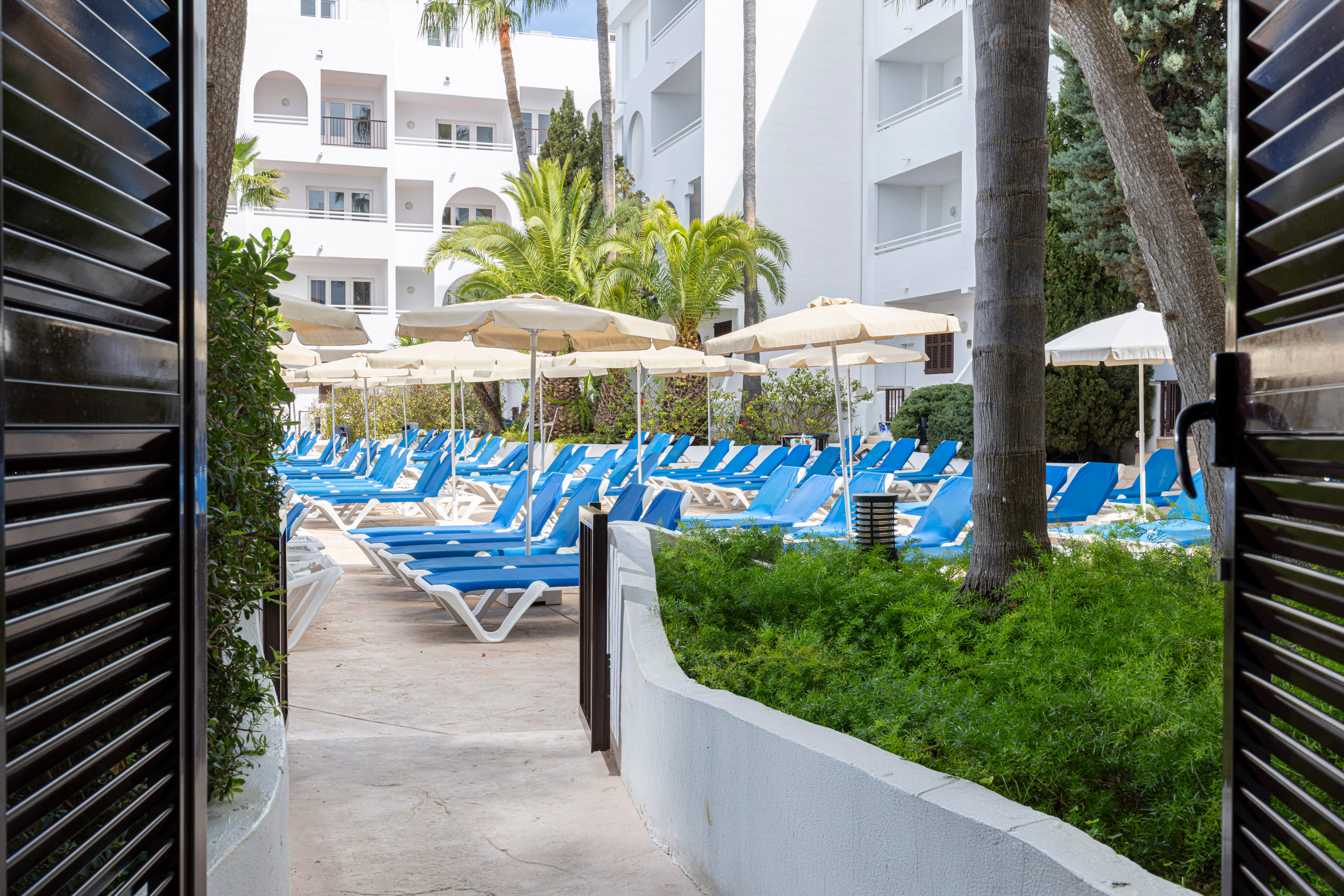 Resort pool area with blue sun loungers, beige umbrellas, and palm trees.