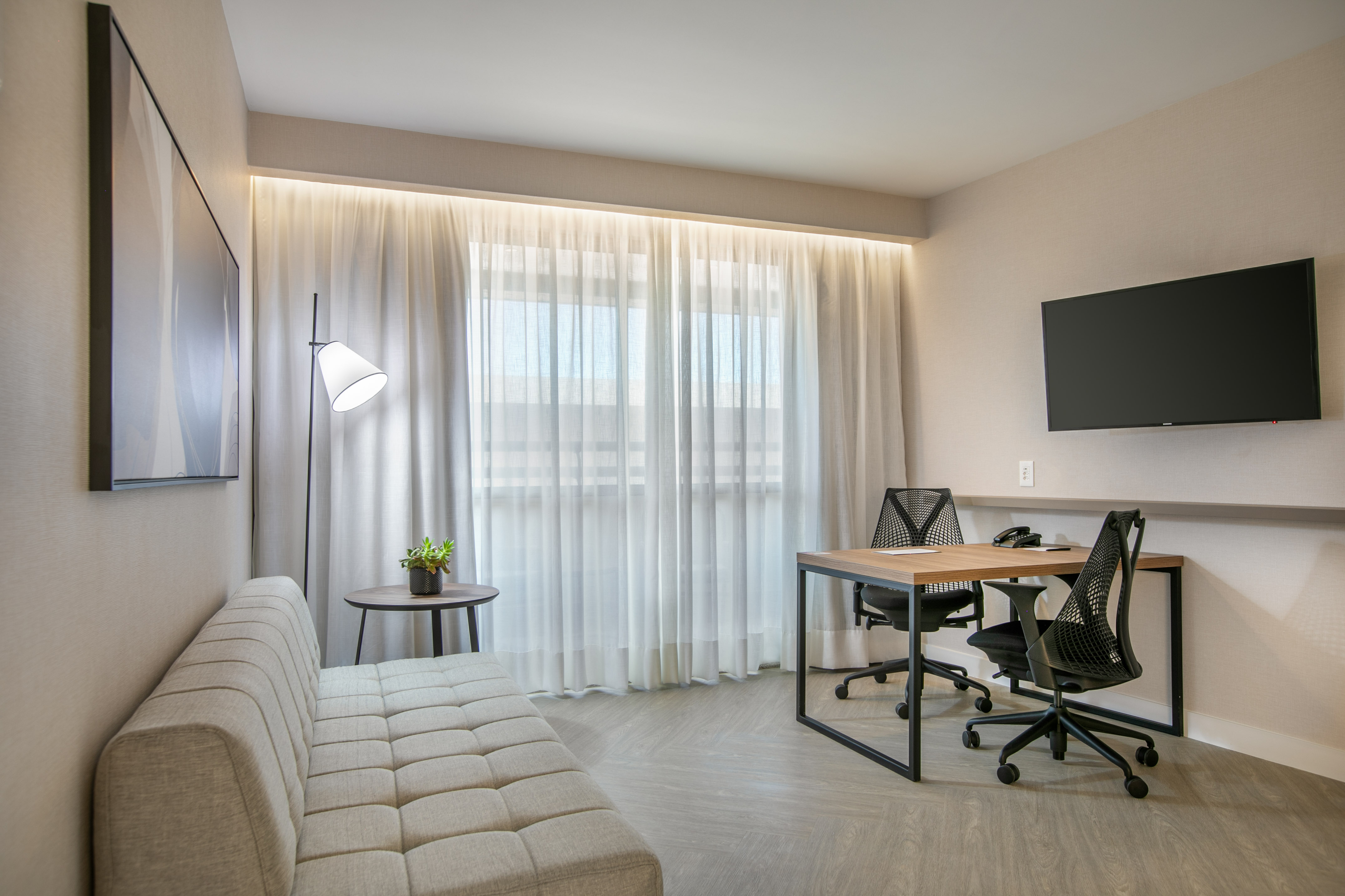 Modern hotel suite with workspace, sofa, and wall-mounted TV.