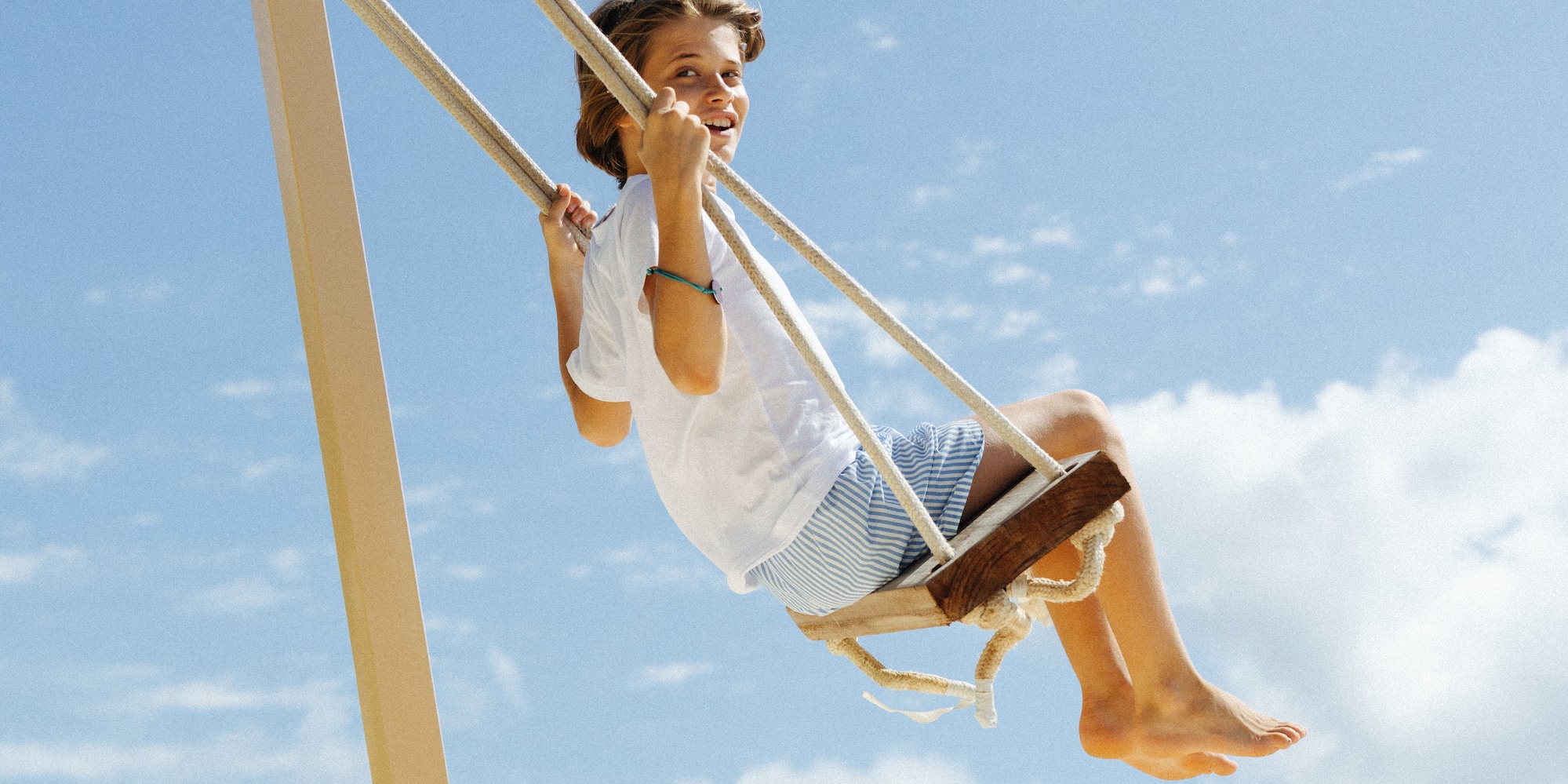 Young boy smiling on a swing at a tropical beach under a blue sky.