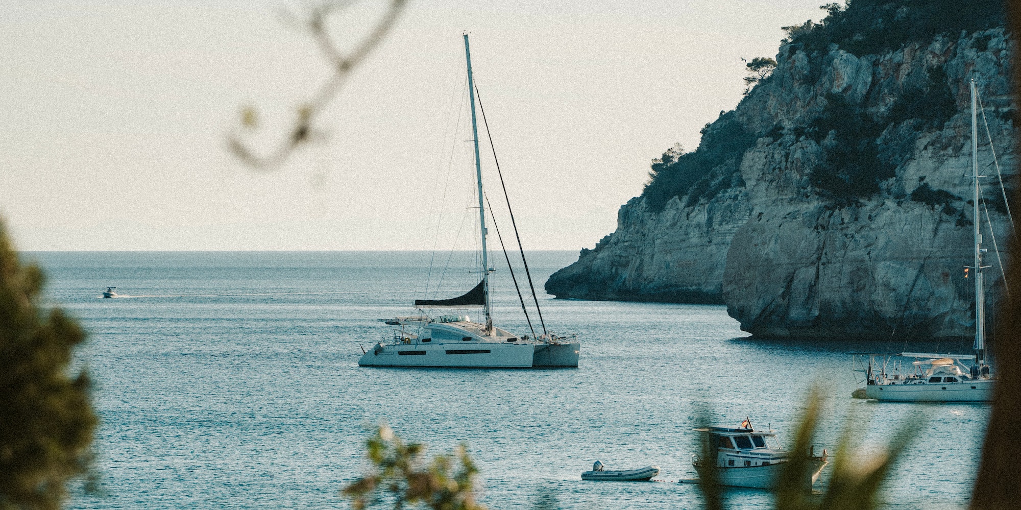 Catamarán blanco en bahía mediterránea con acantilados verdes y mar azul.