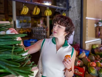 a woman holding an orange and looking at vegetables