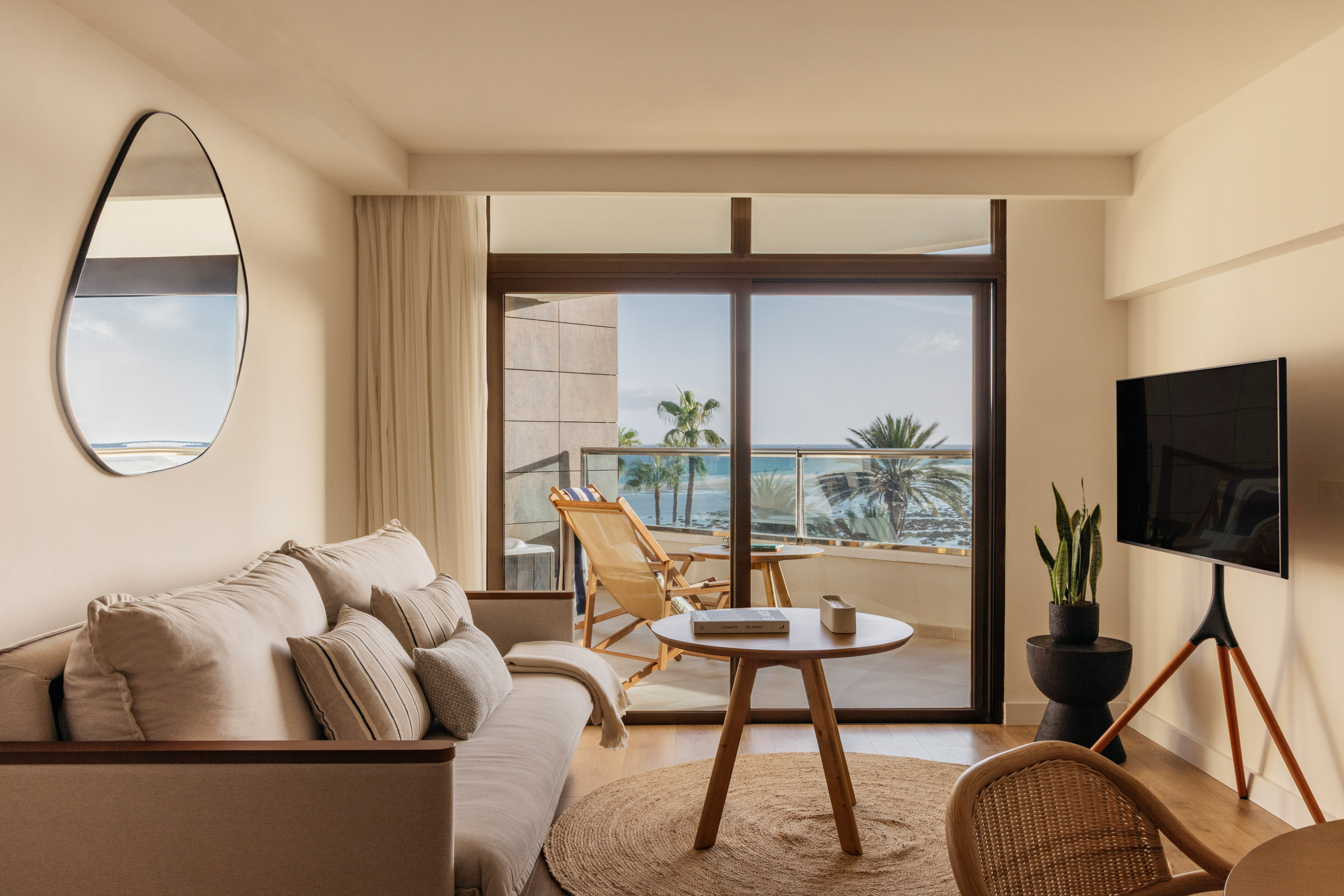 Modern hotel living room with ocean view balcony and palm trees.