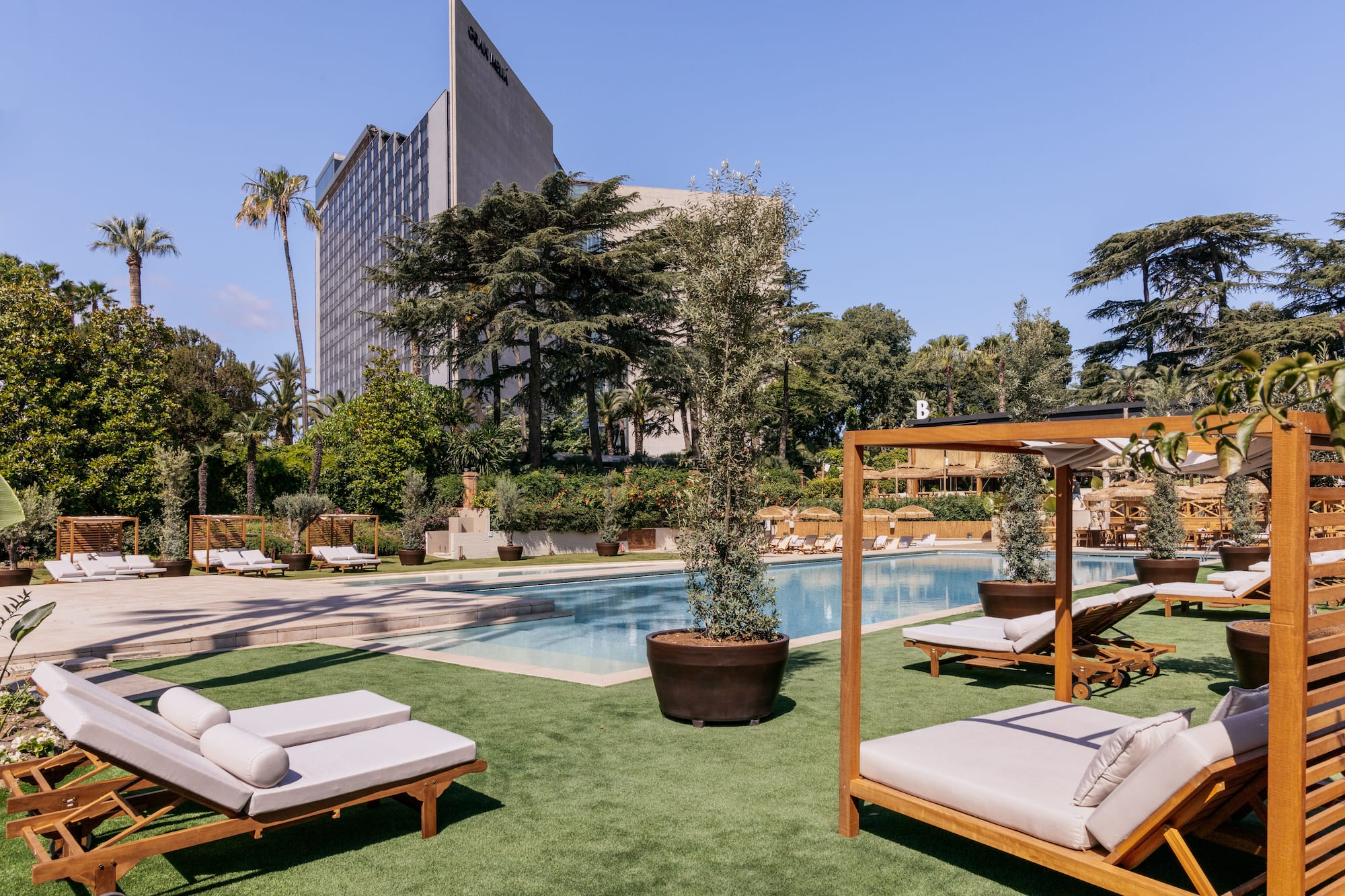 Luxury resort pool with wooden sun loungers, green lawn, and hotel building.