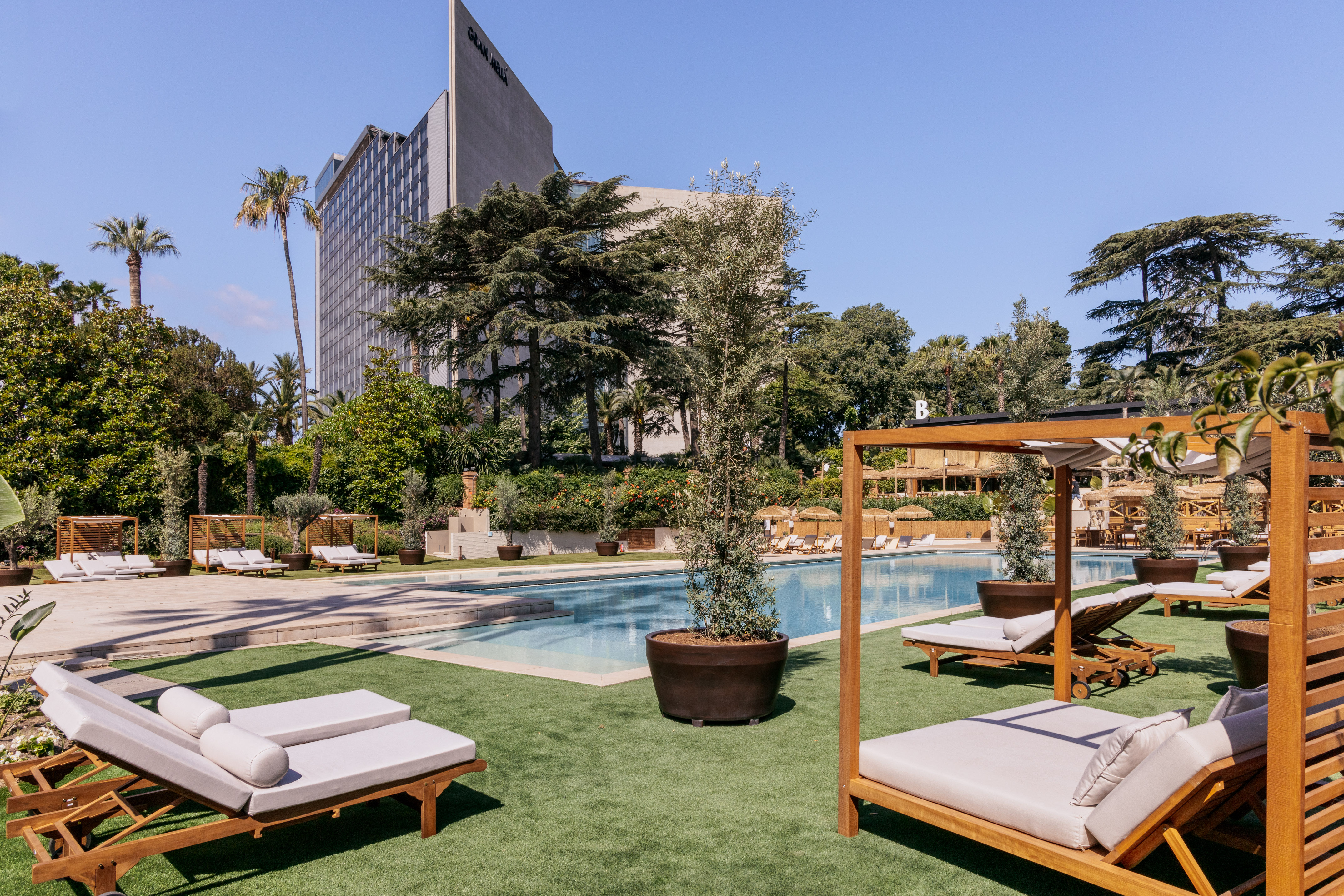 Luxury resort pool with wooden sun loungers, green lawn, and hotel building.