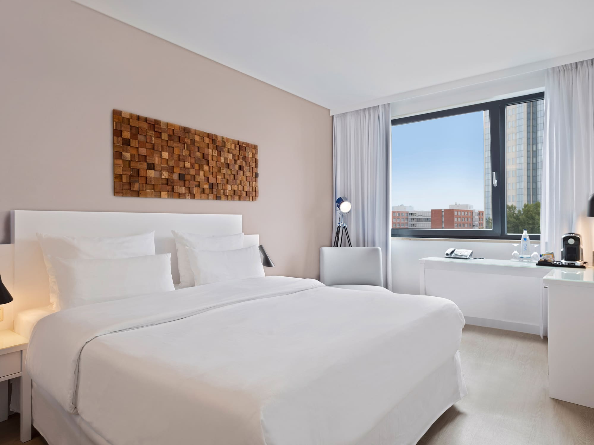 Modern hotel room with white bed, wood wall art, and city view.