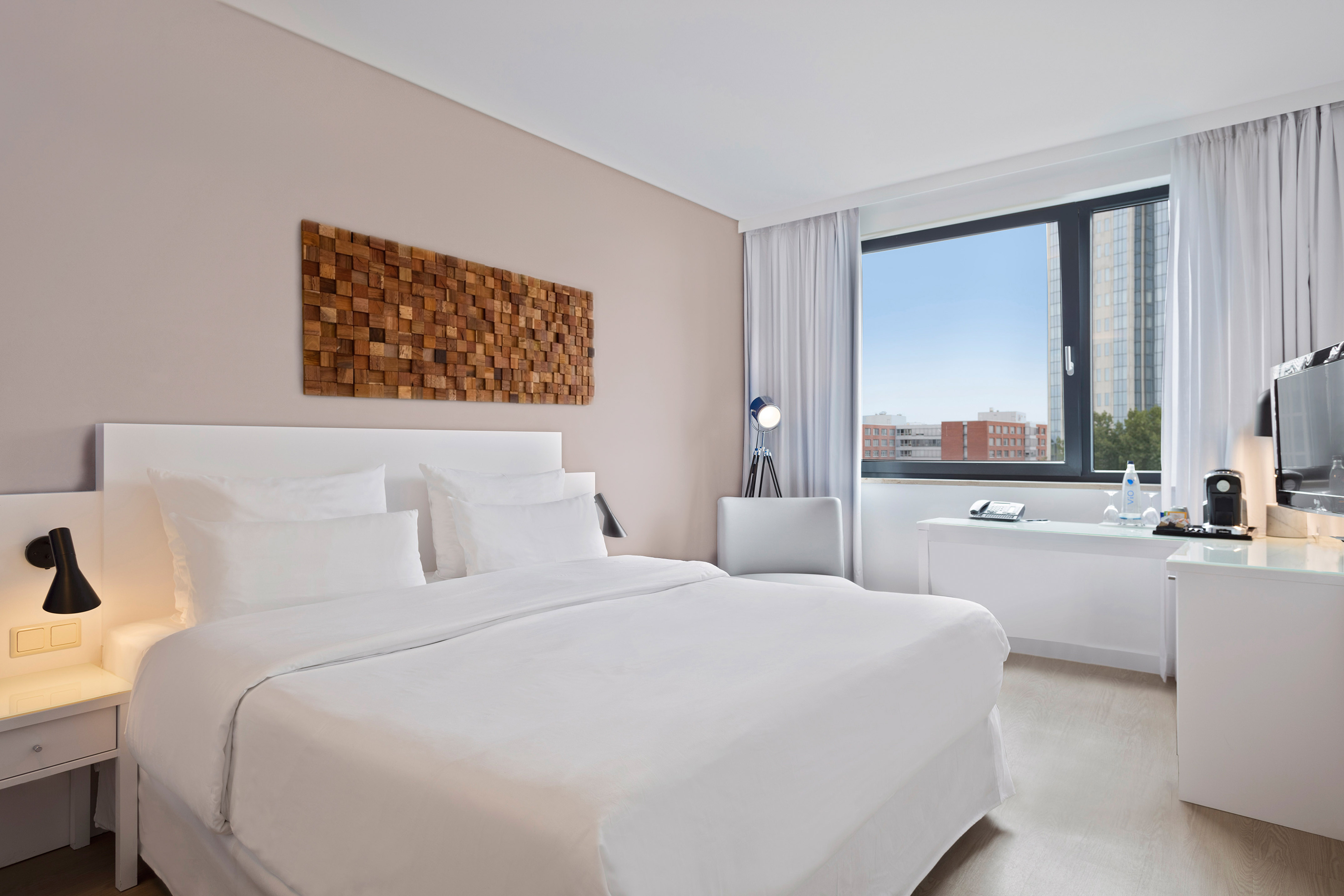 Modern hotel room with white bed, wood wall art, and city view.