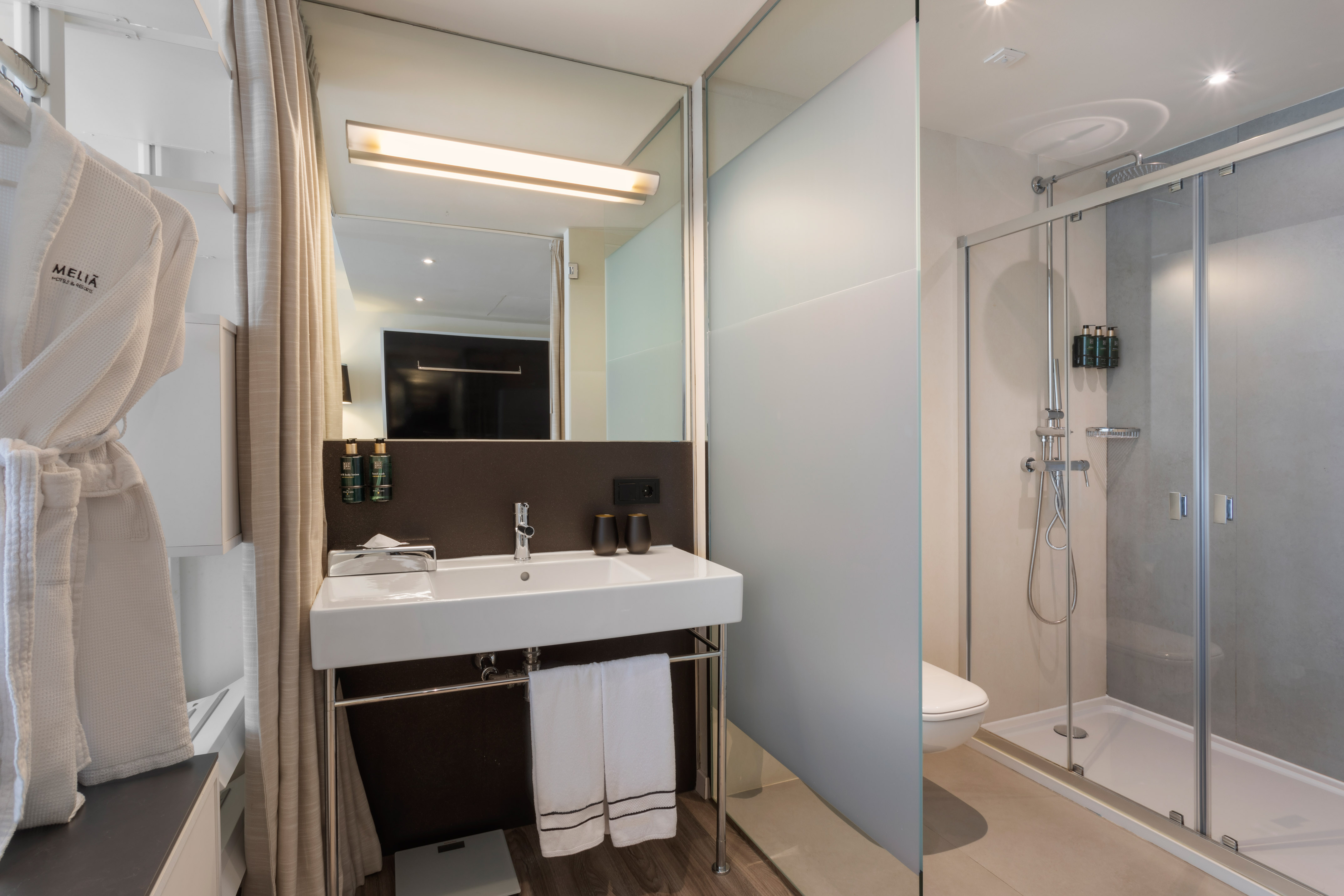 Modern Melia hotel bathroom with vanity, mirror, walk-in shower, and white robe.