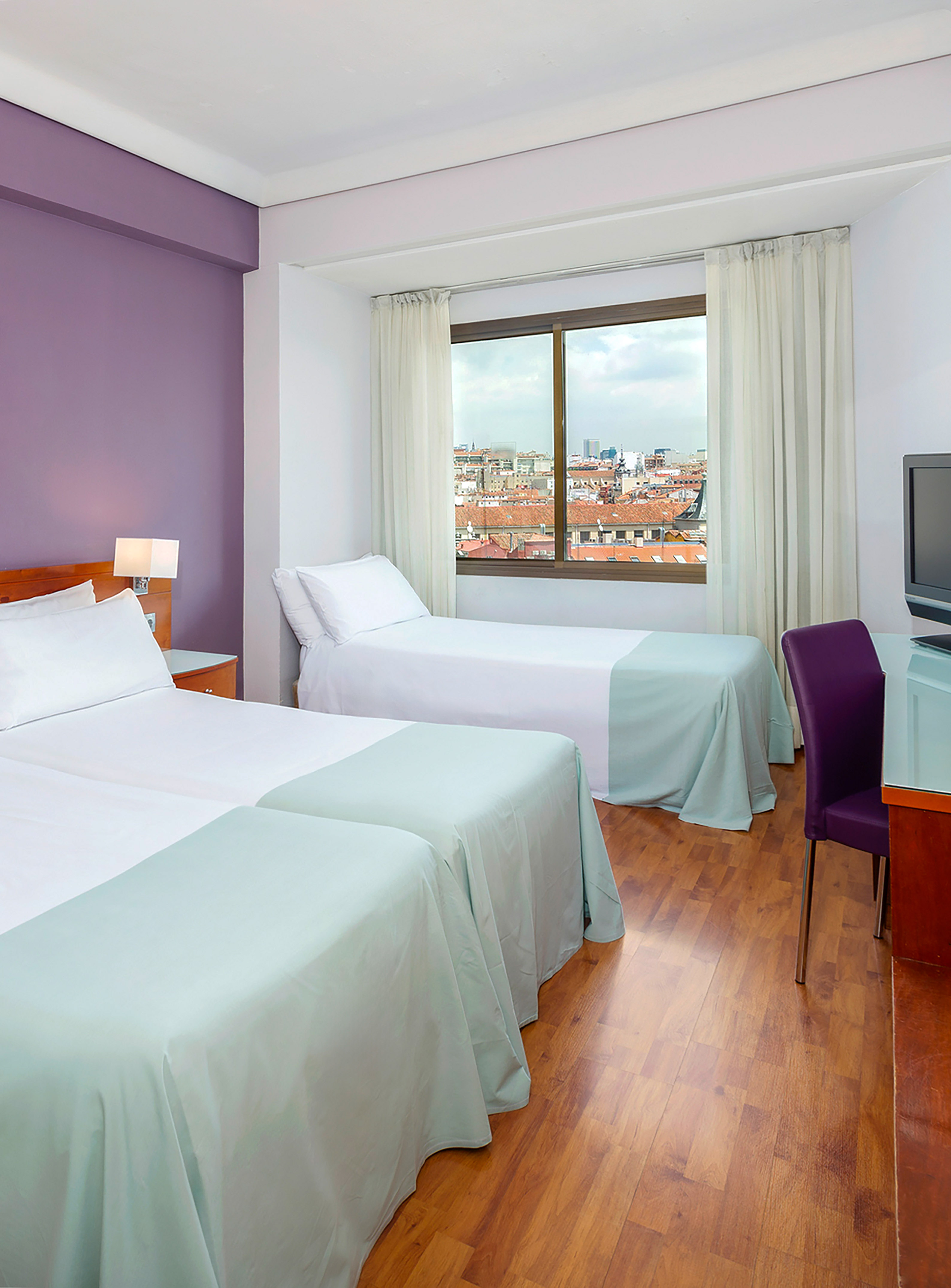 Twin hotel room with city view, purple wall, desk, and wood floor.