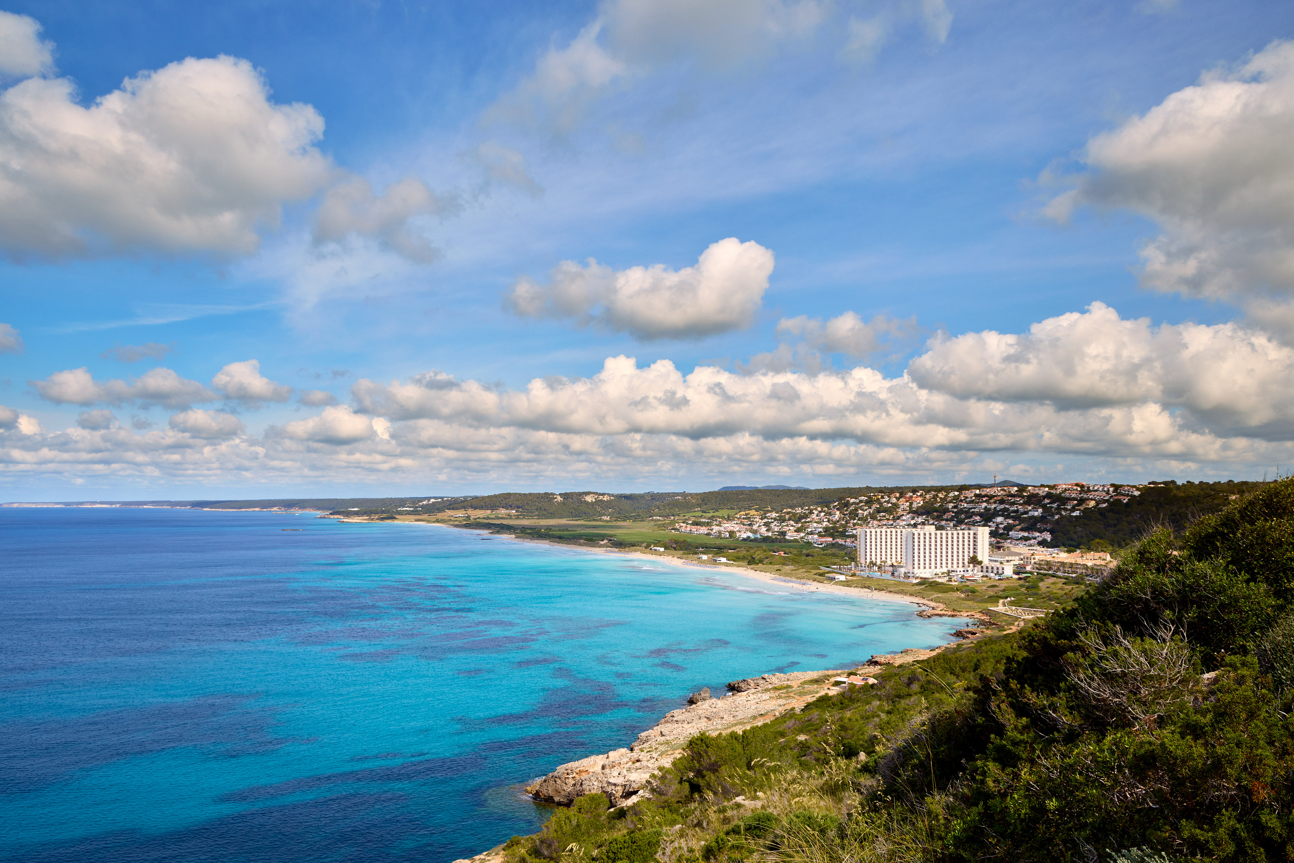 Panoramic turquoise beach, resort hotel, and coastal town under blue sky.