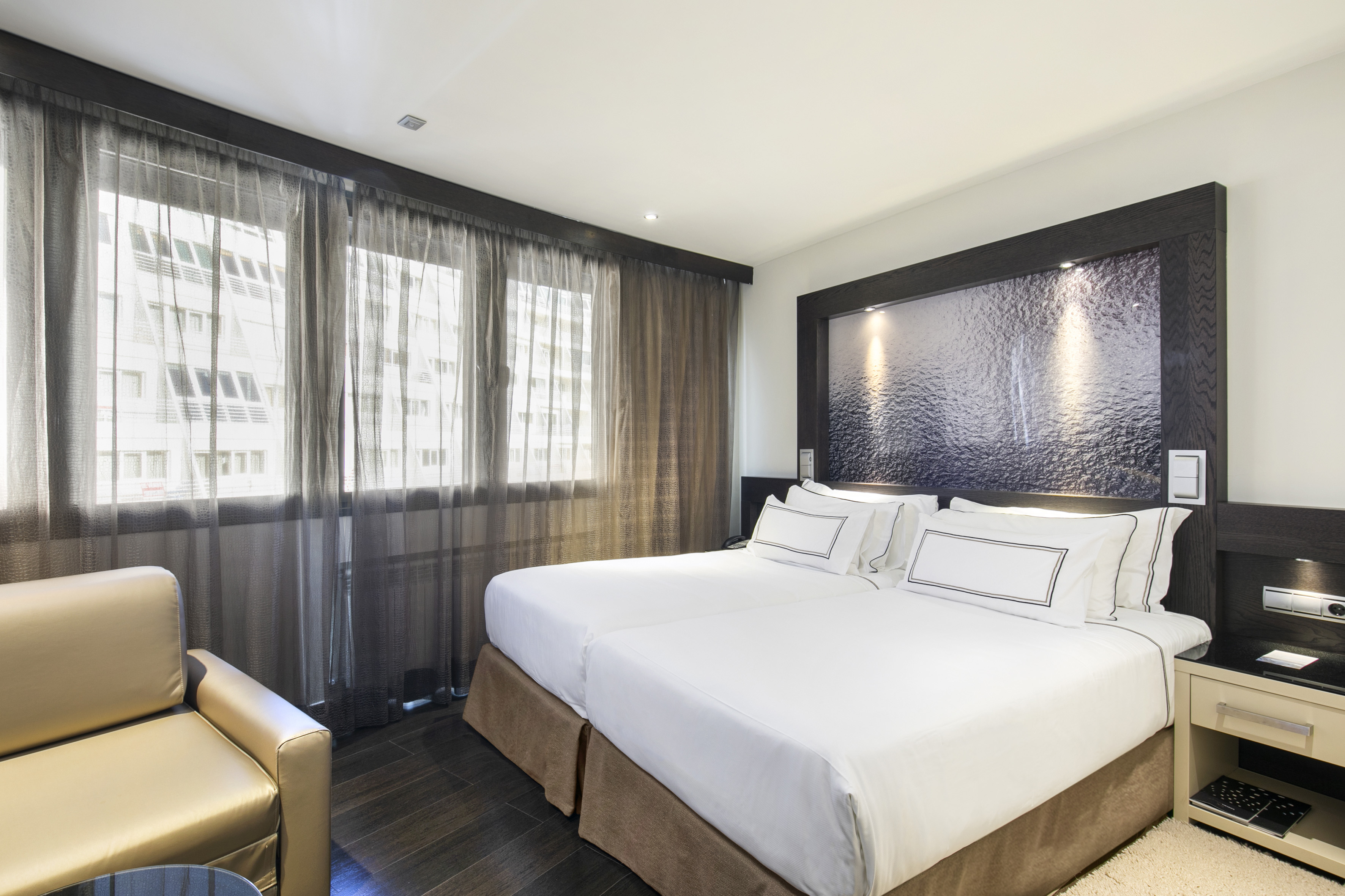 Modern hotel room with twin beds, dark wood accents, and sheer curtains.