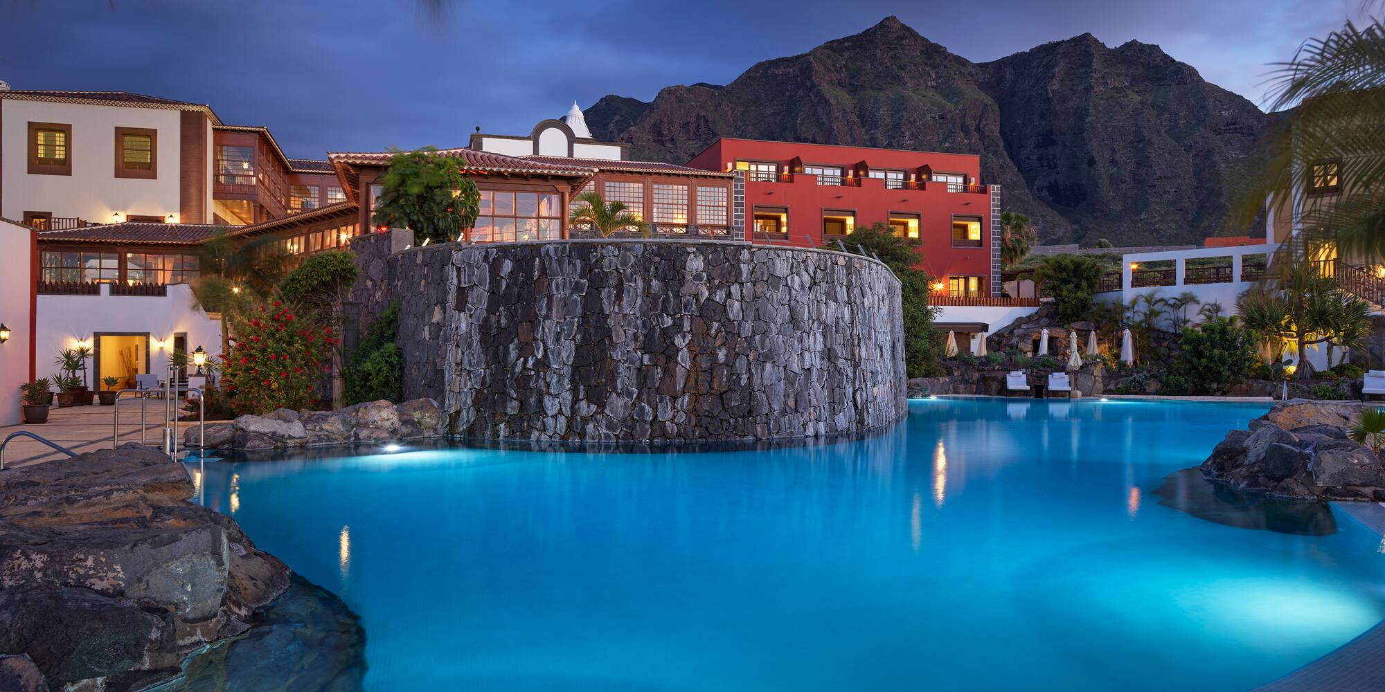 Illuminated resort pool, stone facade, and mountain backdrop at dusk.