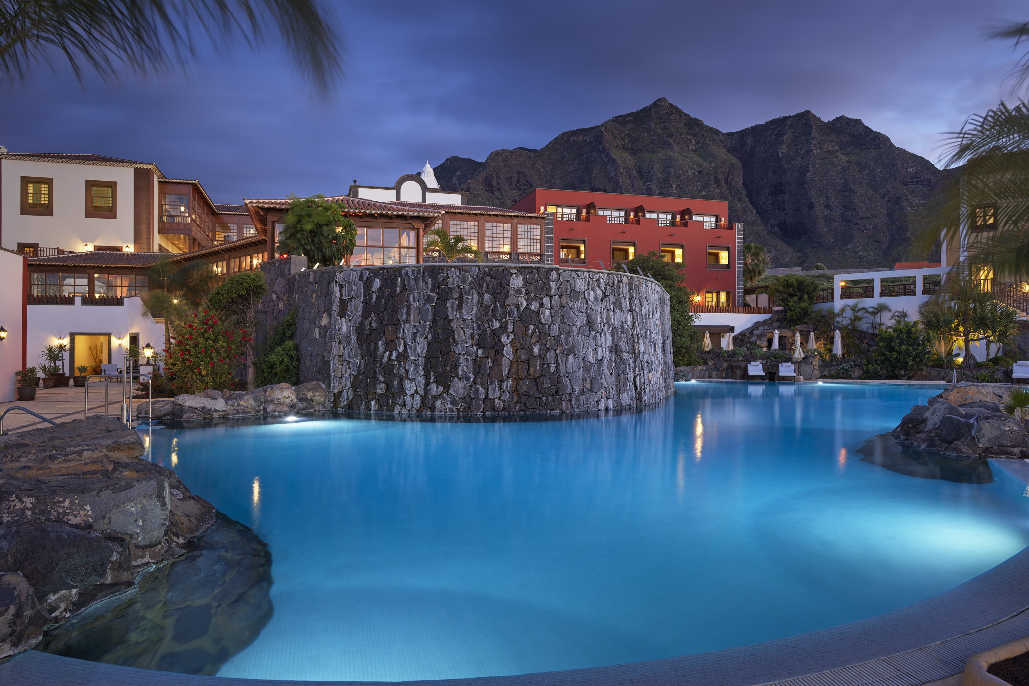 Illuminated resort pool, stone facade, and mountain backdrop at dusk.