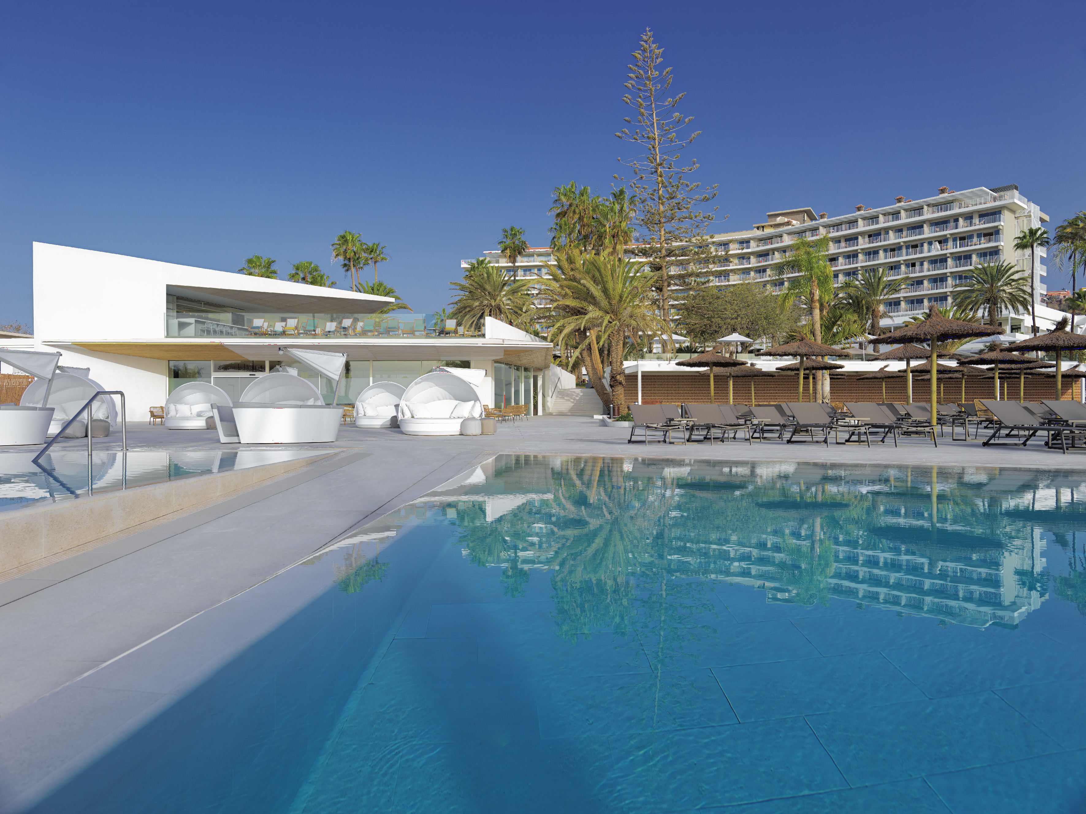 Modern resort pool area with luxury hotel, palm trees, and white cabanas.