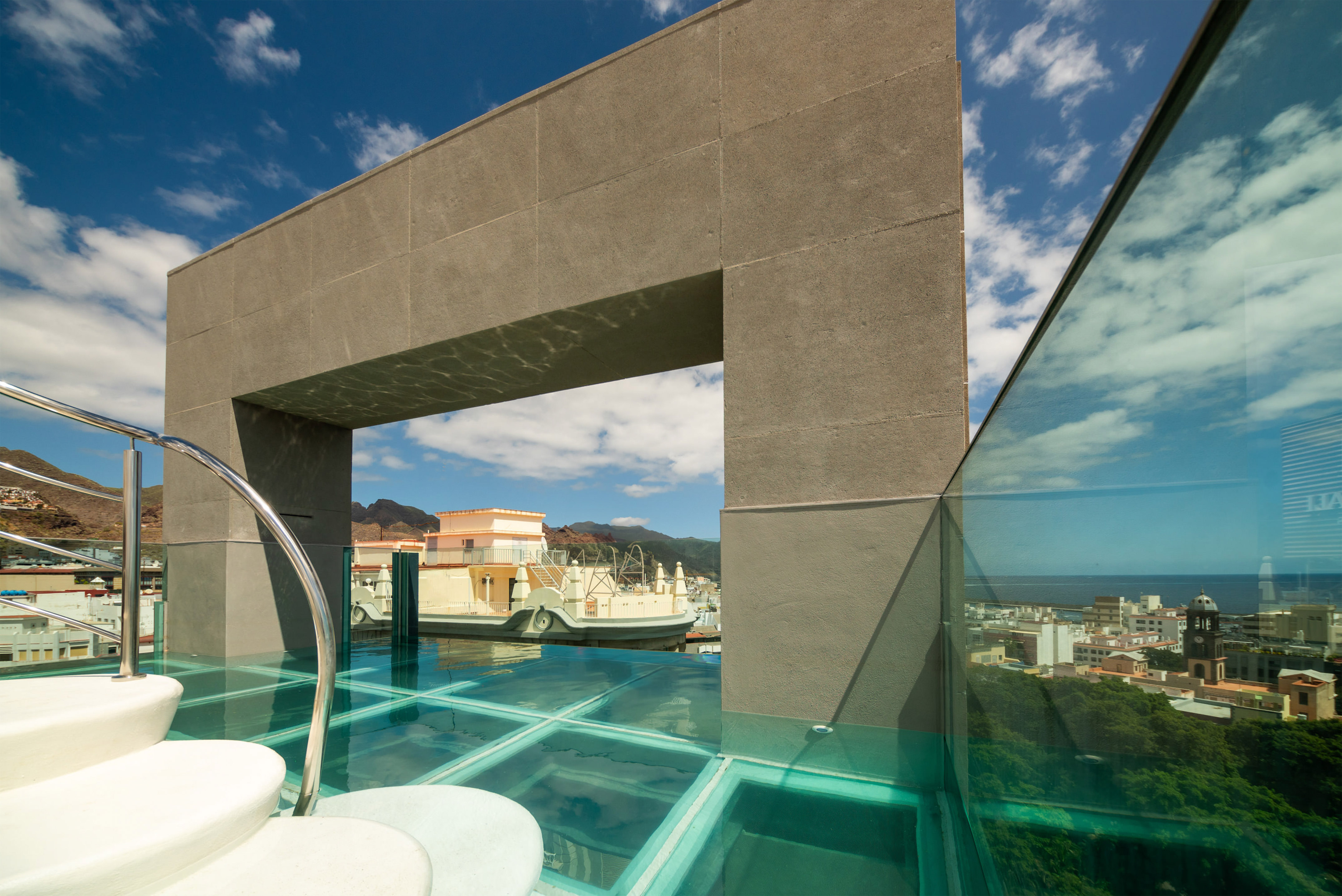 Rooftop glass-bottom pool with city, mountain, and ocean views under blue sky.
