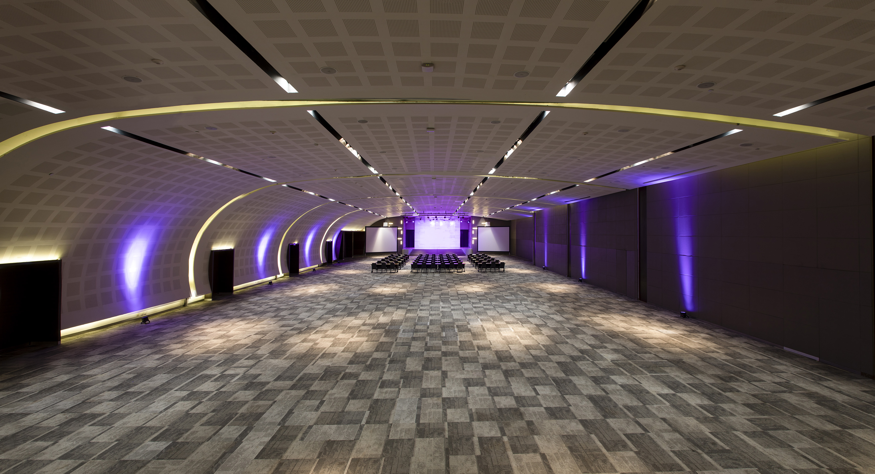 Modern event hall with stage, empty seating, and purple wall uplighting.