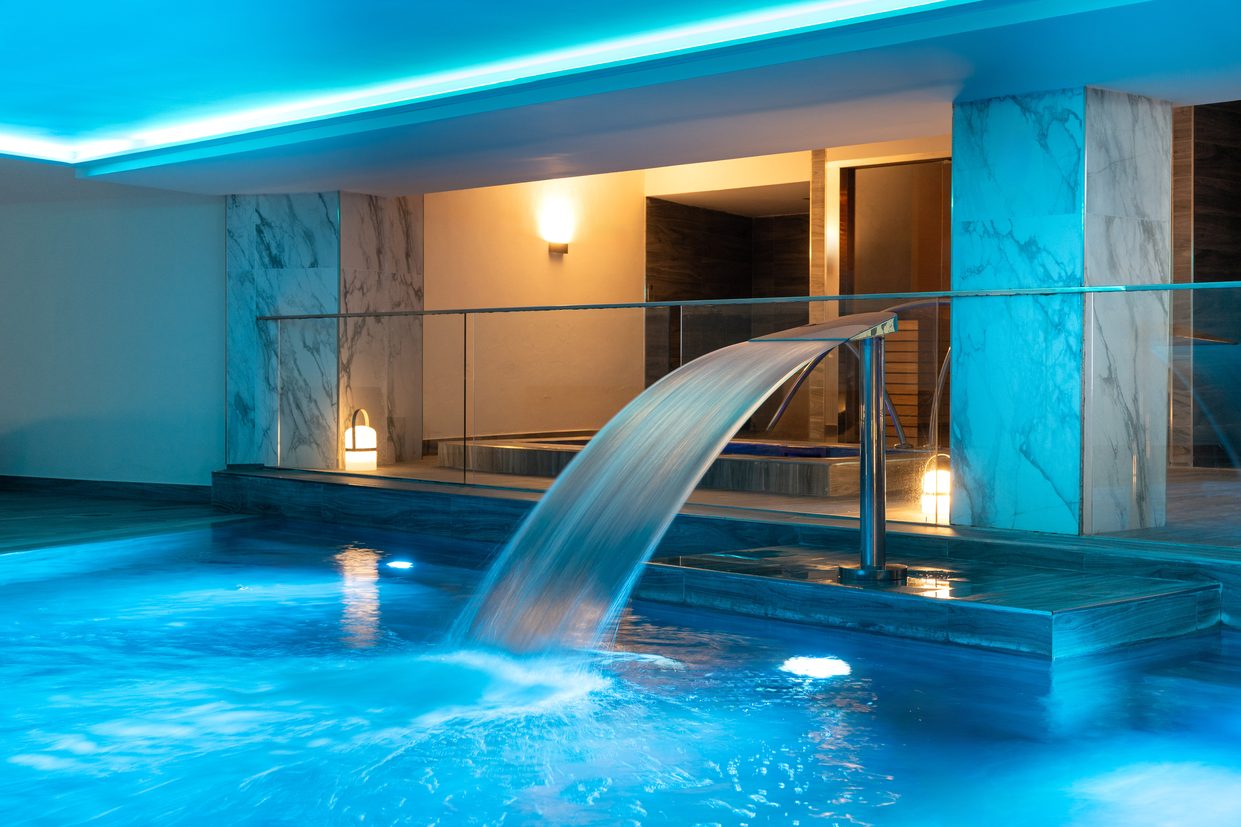Modern indoor spa pool with blue lighting and cascading waterfall.