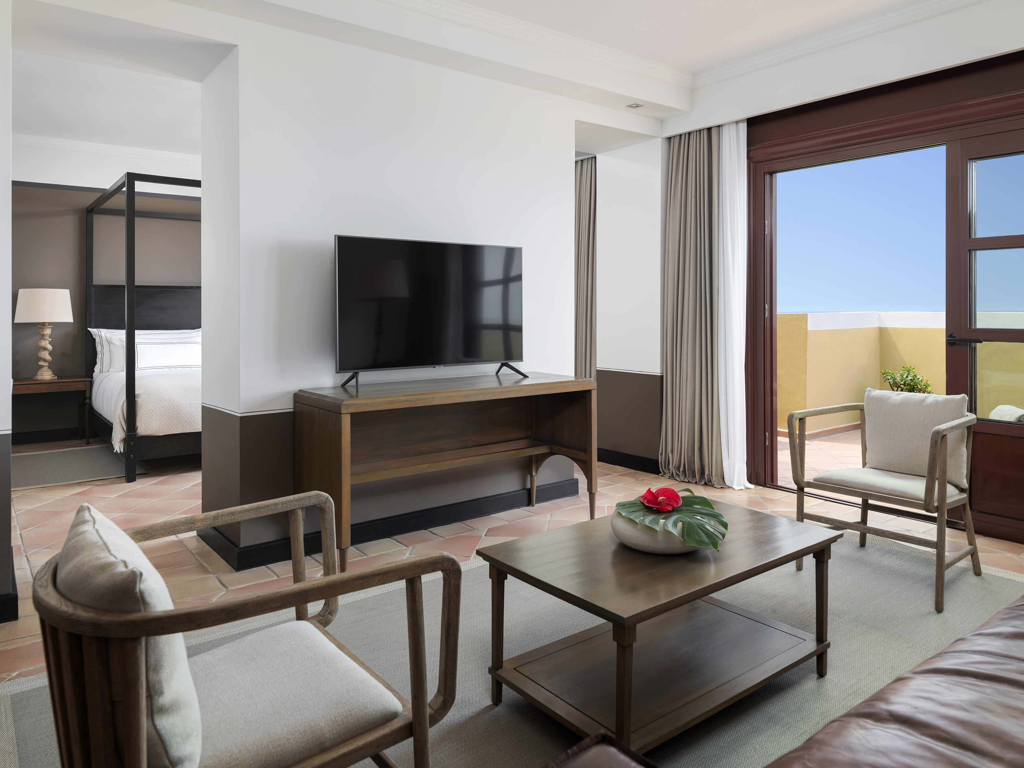 Hotel suite: living room with TV, connected bedroom, and sunny balcony.