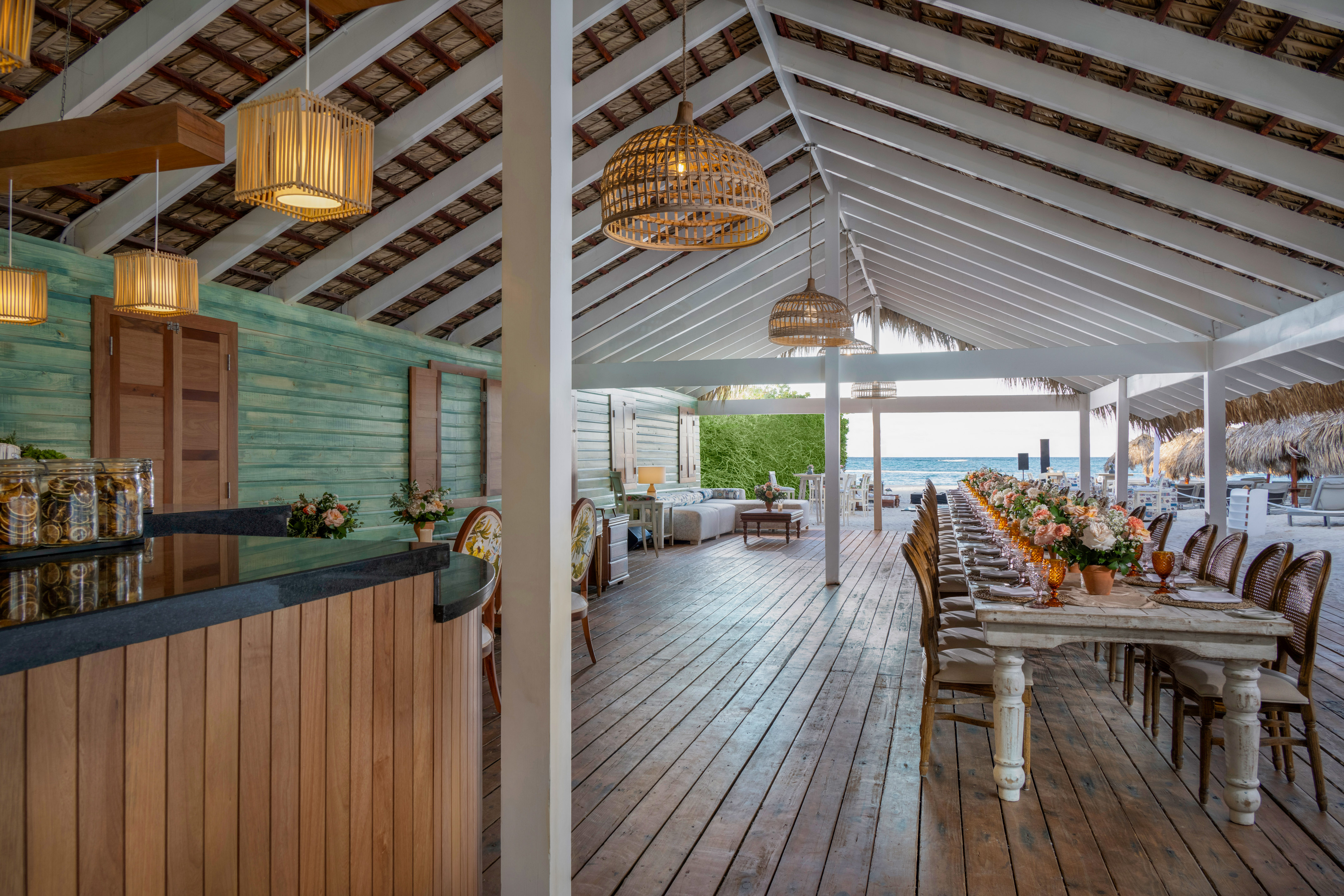 Beachfront dining pavilion with long table, flowers, ocean view, and bar area.