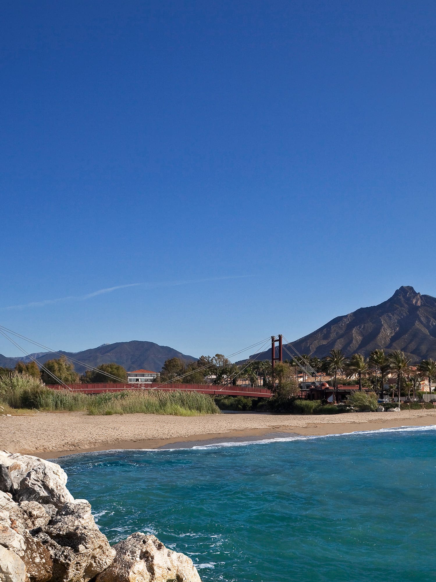 Marbella beach, Puente Romano red bridge, white hotel, and mountains under blue sky.