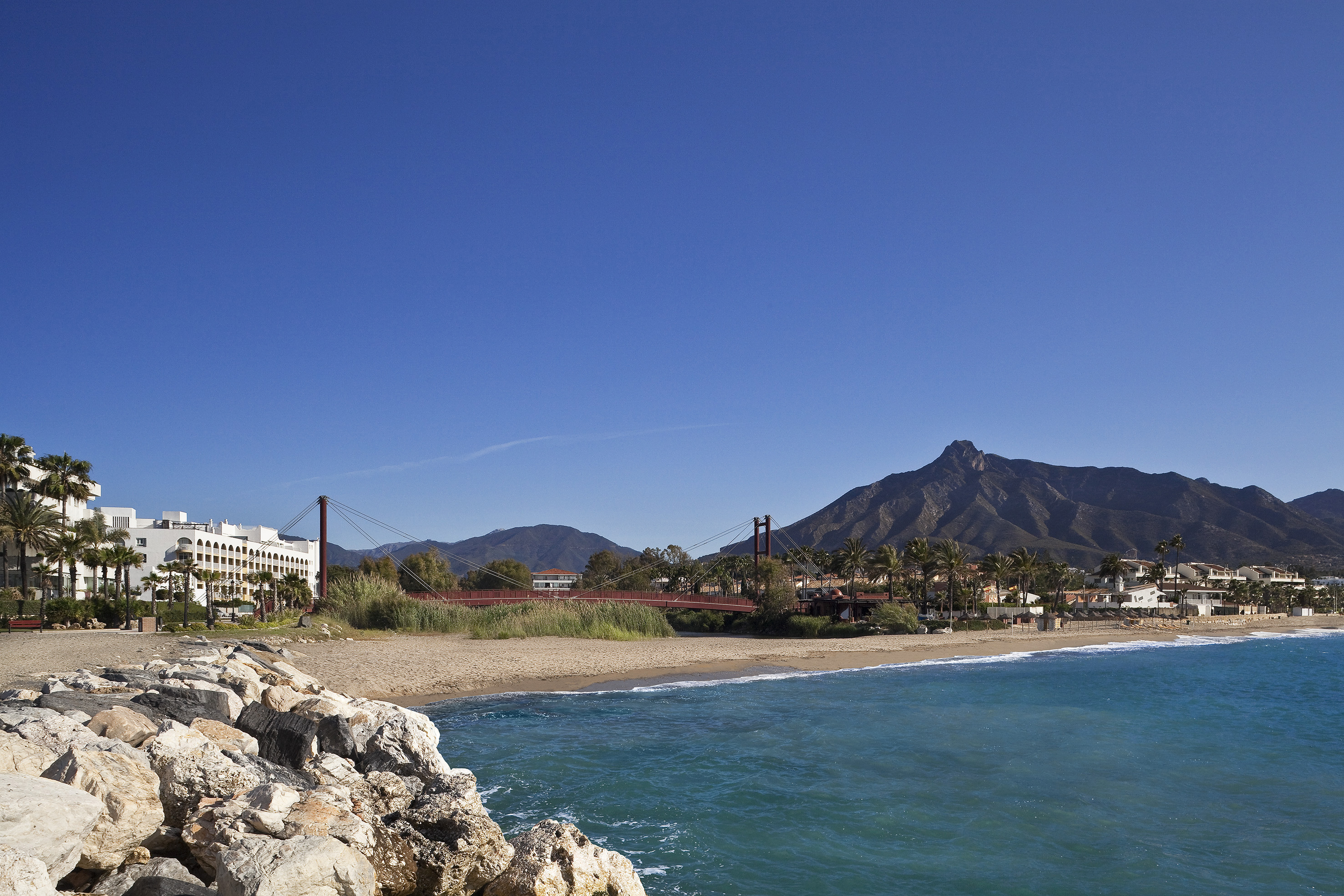 Marbella beach, Puente Romano red bridge, white hotel, and mountains under blue sky.
