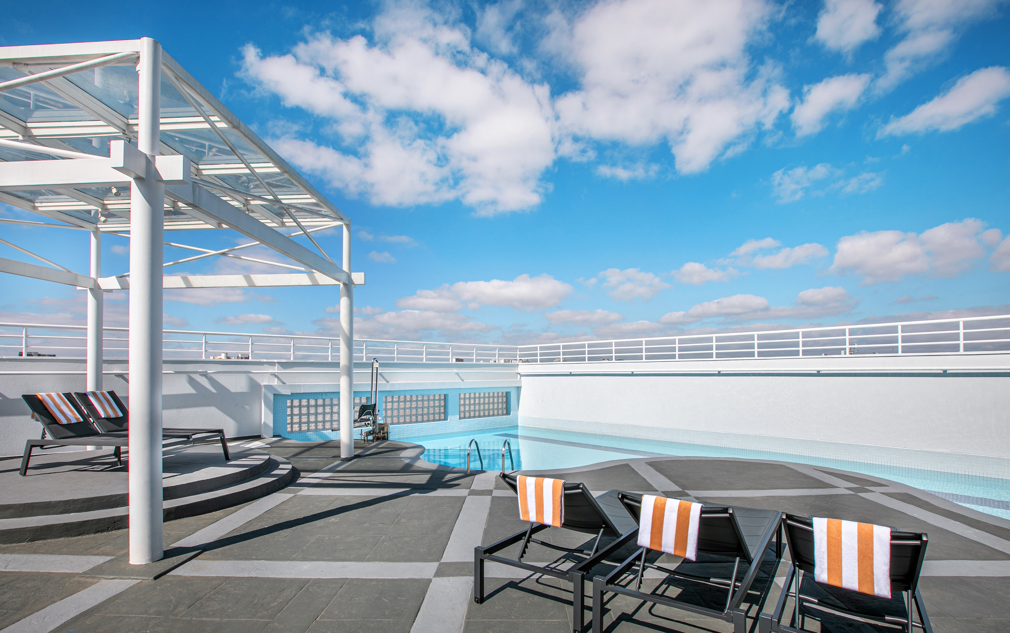 Modern rooftop pool, white pergola, lounge chairs, orange towels, blue sky.