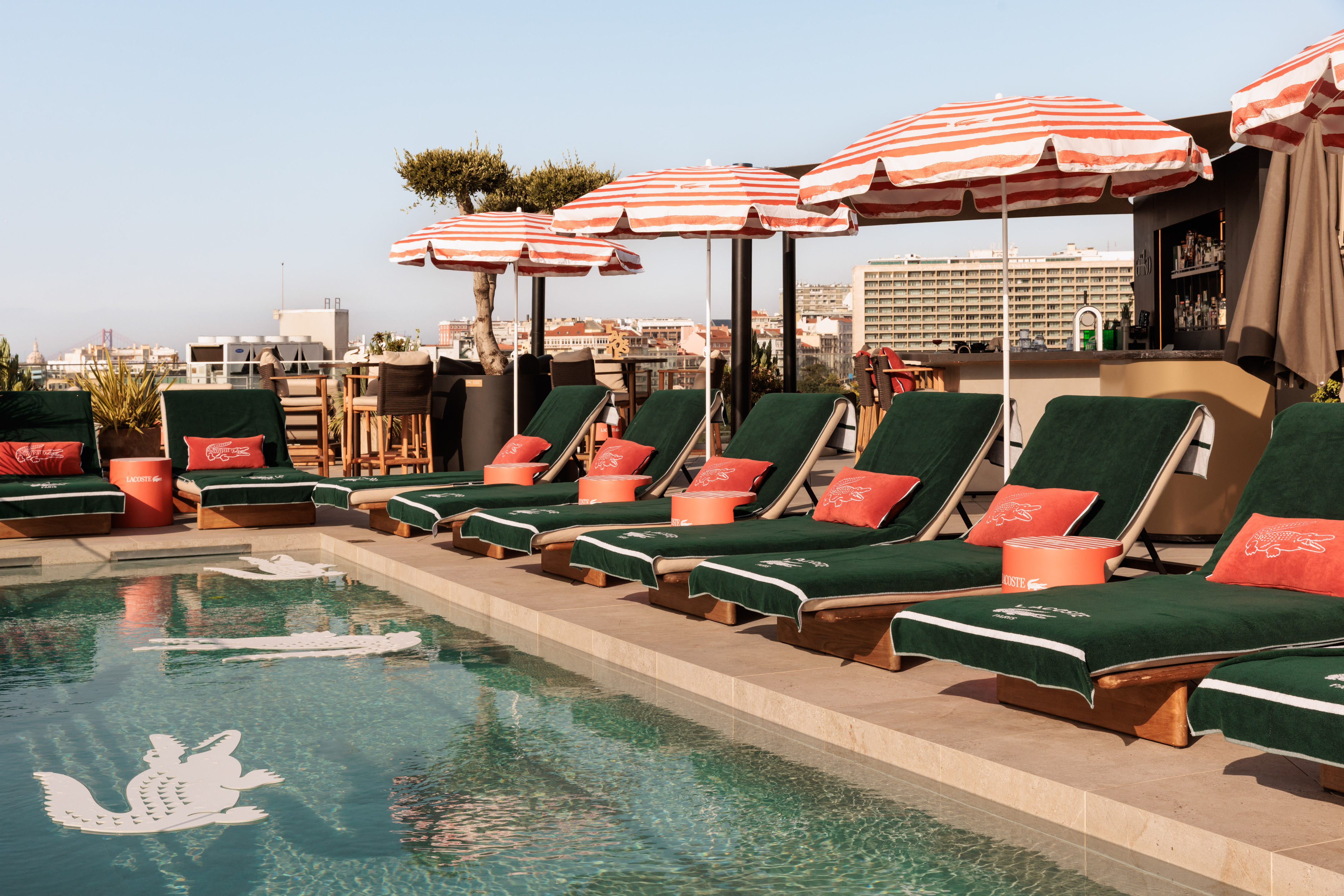 Rooftop pool with green Lacoste lounge chairs, striped umbrellas, and city skyline.