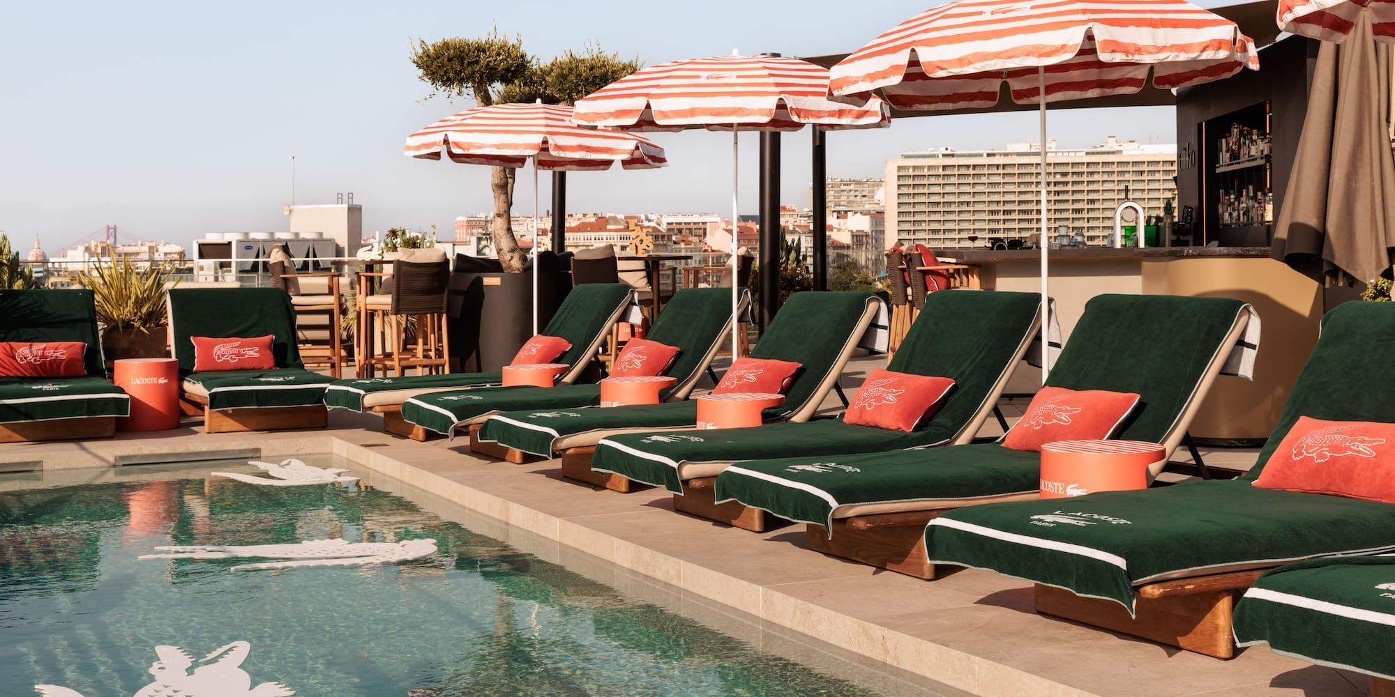 Lacoste rooftop pool, green loungers, red umbrellas, and city skyline.