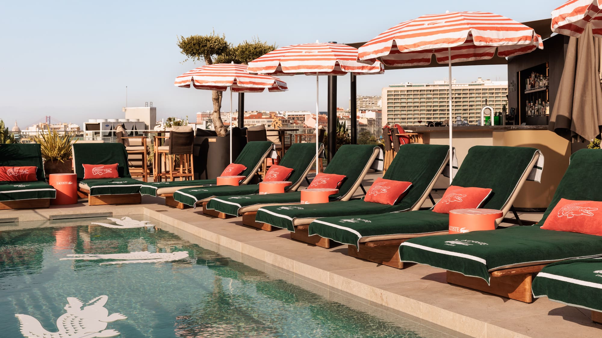 Lacoste rooftop pool, green loungers, red umbrellas, and city skyline.