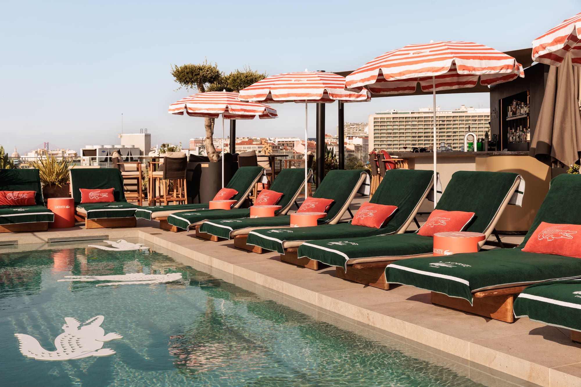 Rooftop pool with green Lacoste lounge chairs, striped umbrellas, and city skyline.