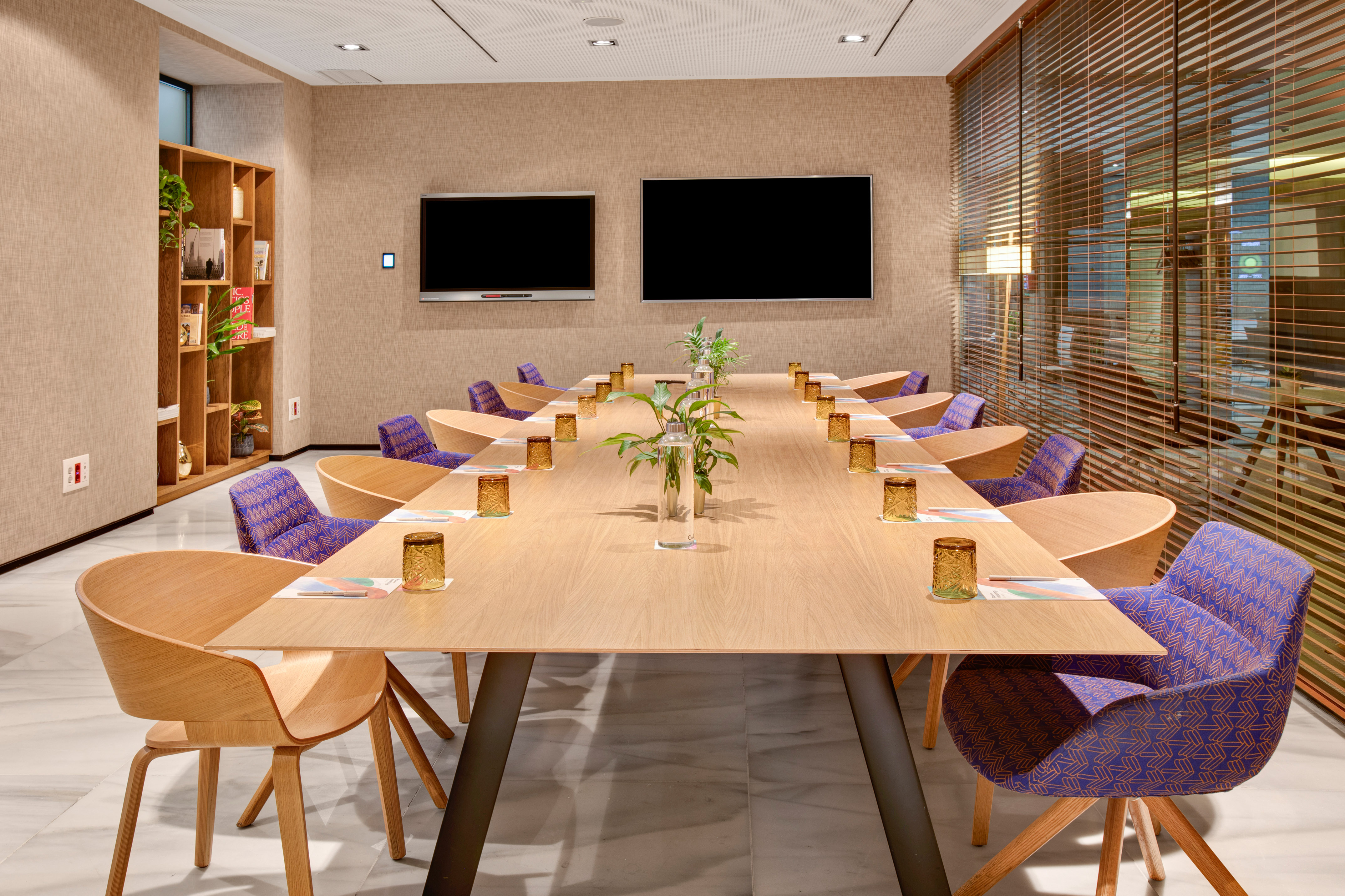 Modern conference room with long wooden table, purple chairs, and dual screens.