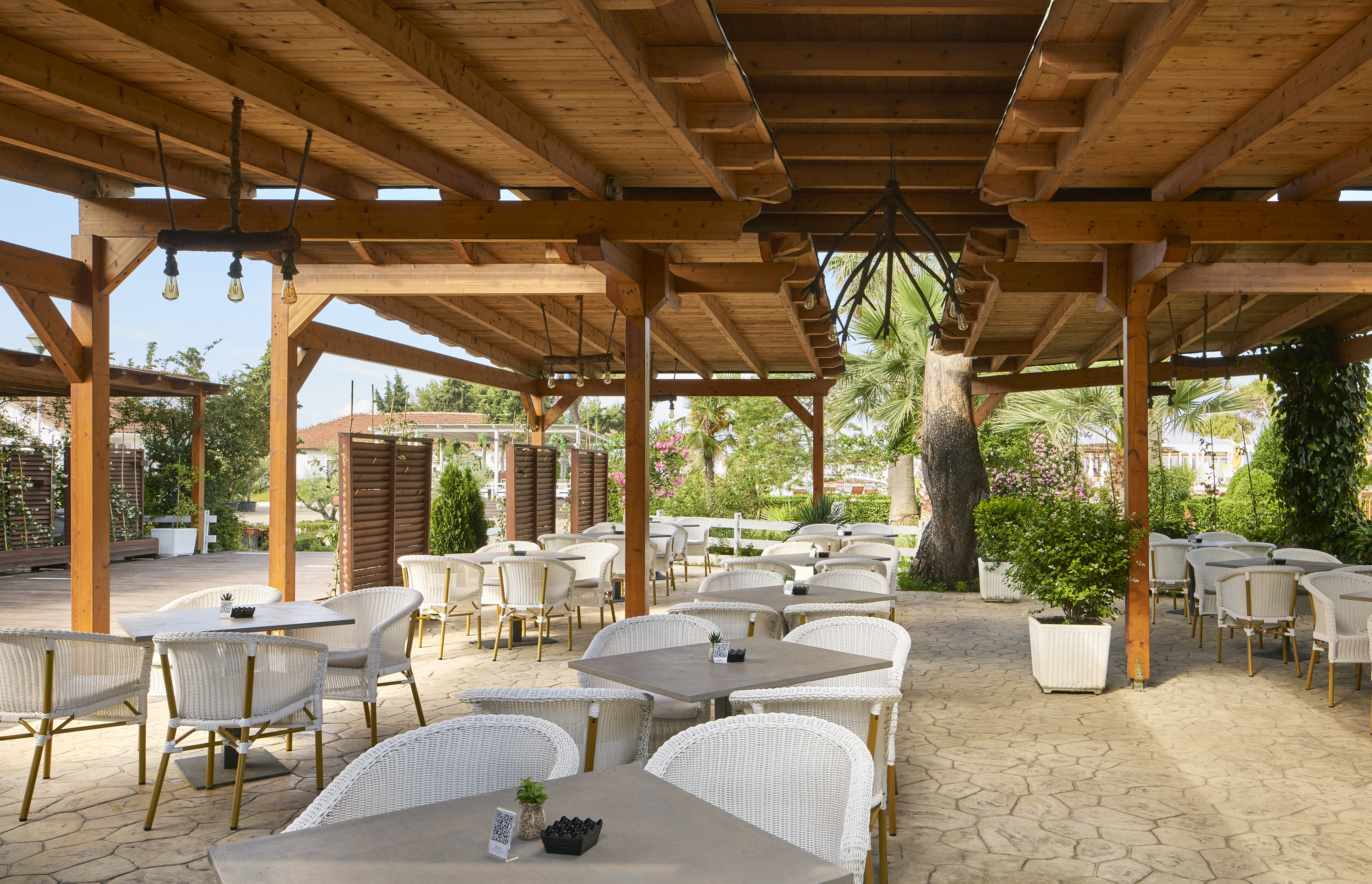 Shaded outdoor dining patio with wooden pergola, wicker seating, and lush plants.