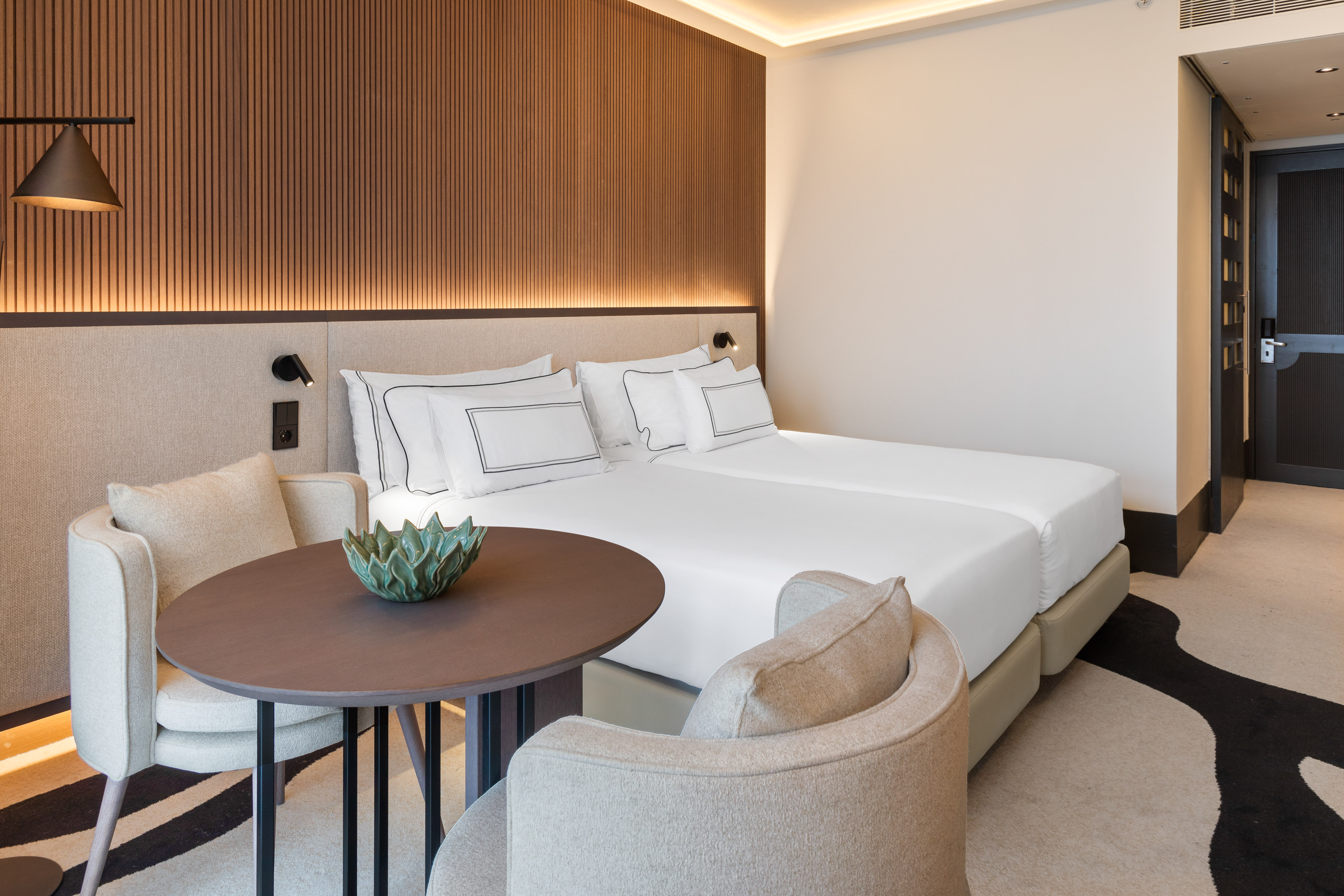 Contemporary hotel room with twin beds, wood paneling, and seating area.