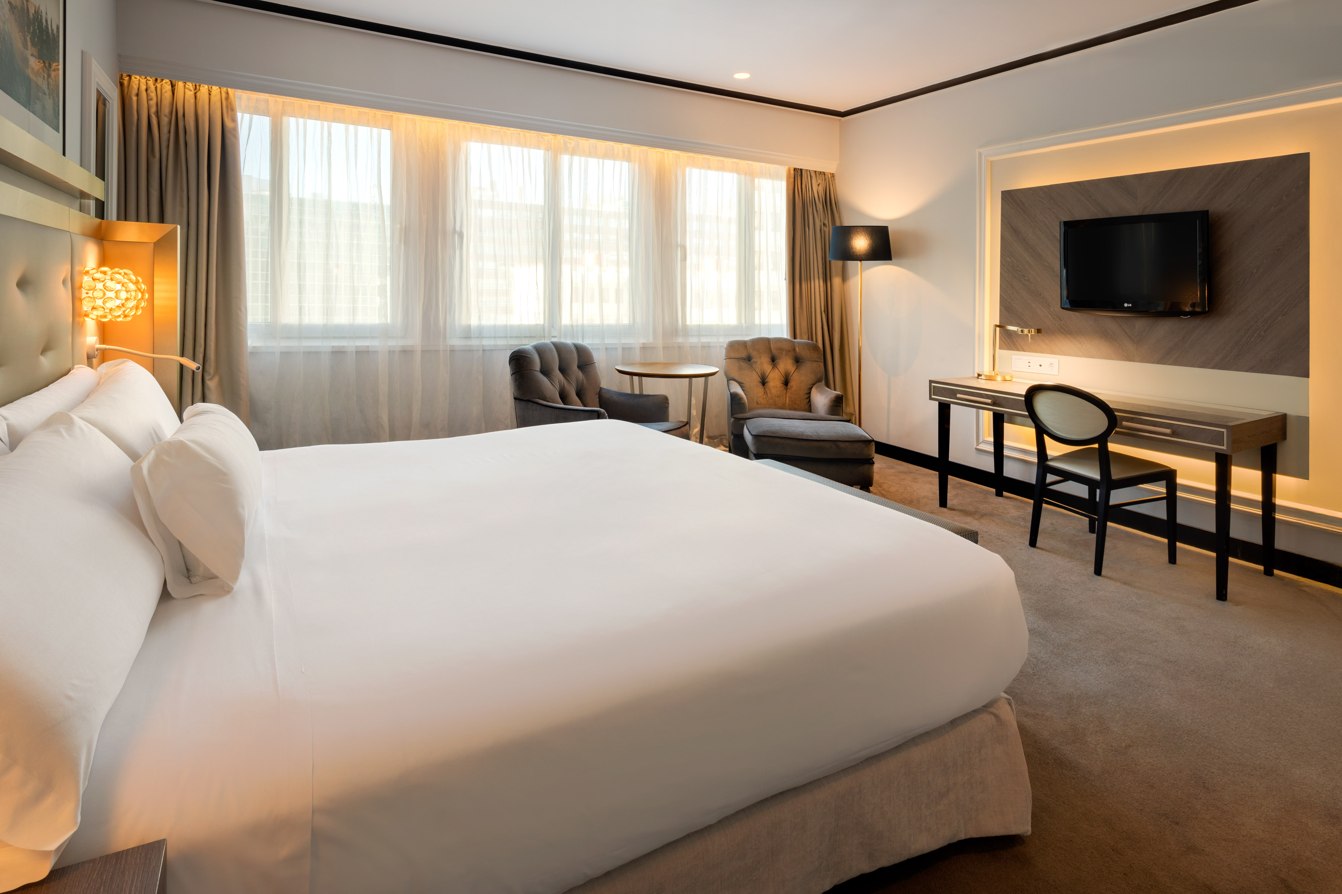 Modern hotel room with king bed, desk, sitting area, and bright window.