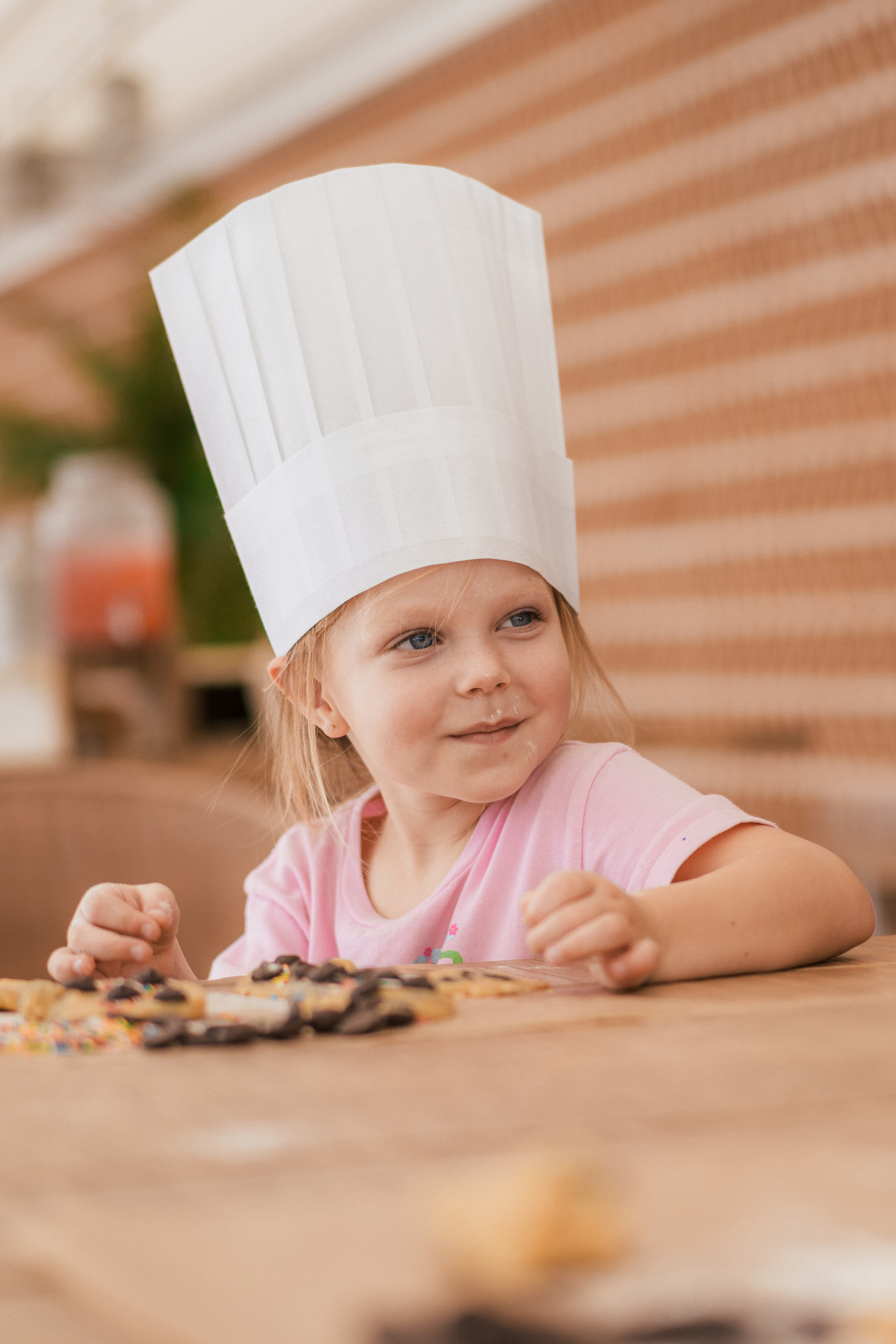 Young girl in chef hat decorating cookies with sprinkles on wooden table.