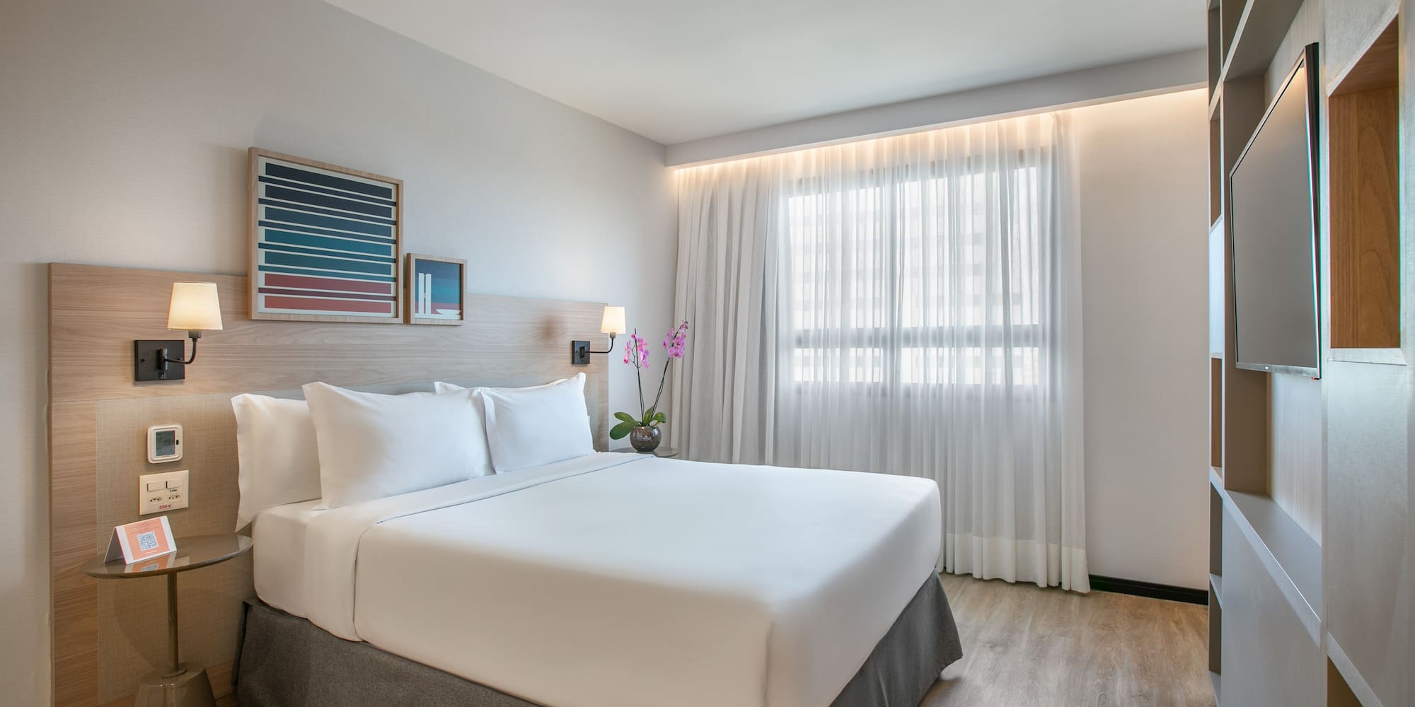 Modern hotel room with king bed, white bedding, wood accents, and window.
