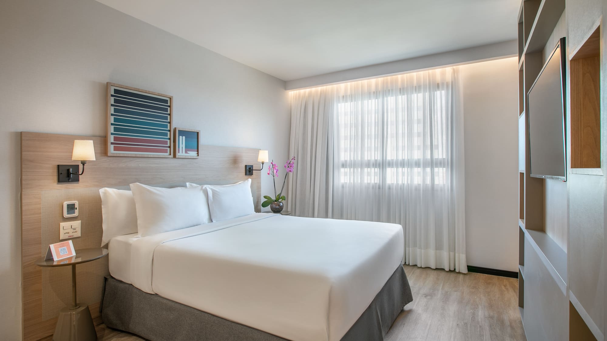 Modern hotel room with king bed, white bedding, wood accents, and window.