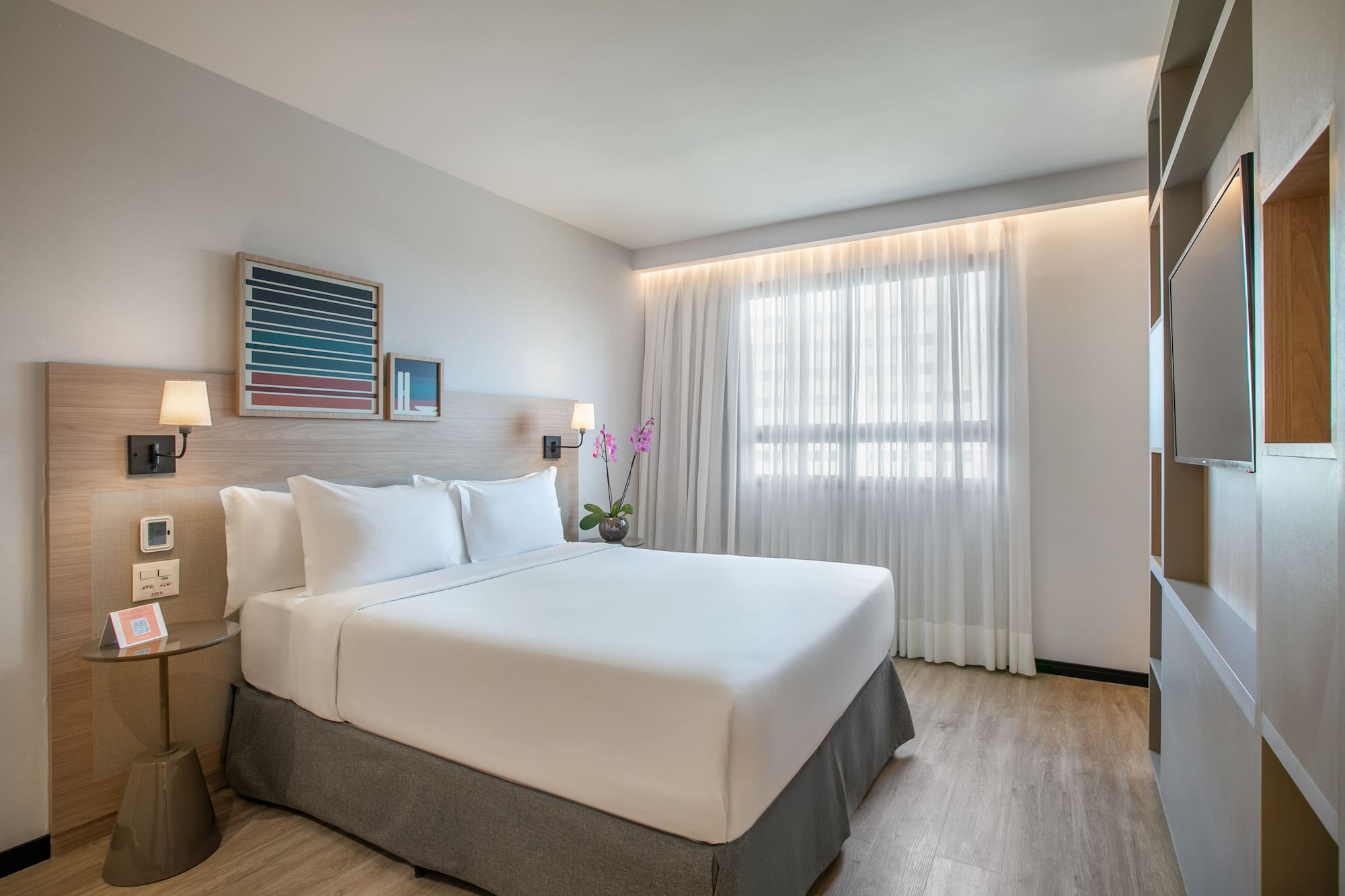 Modern hotel room with king bed, white bedding, wood accents, and window.