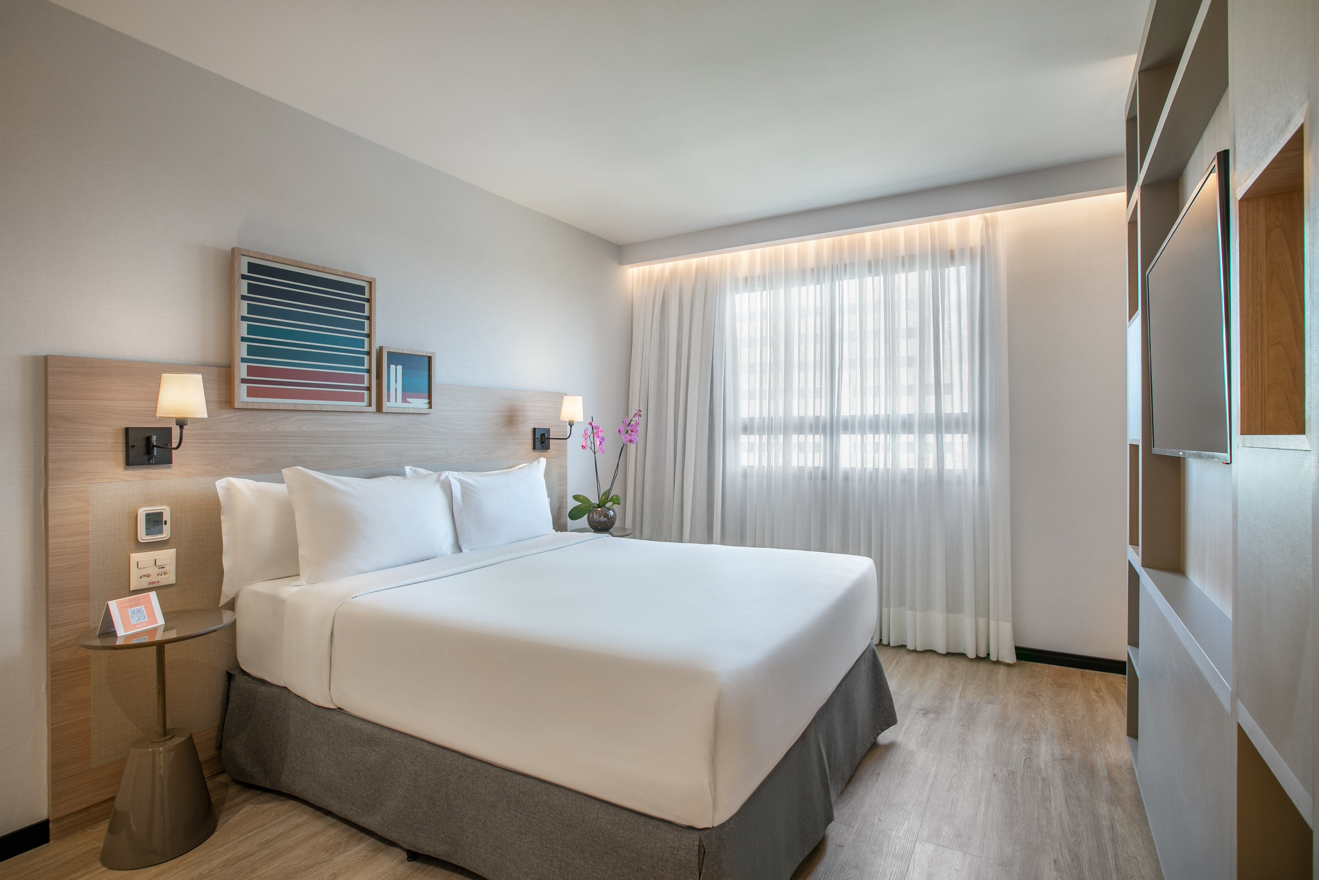Modern hotel room with king bed, white bedding, wood accents, and window.