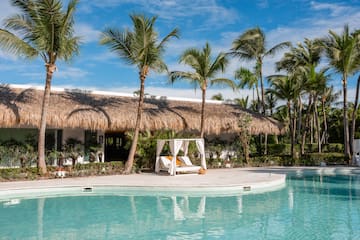 Tropical resort pool with palm trees, thatched building, and poolside cabana.