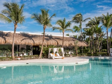Turquoise resort pool with palm trees, thatched roof, and white cabana.
