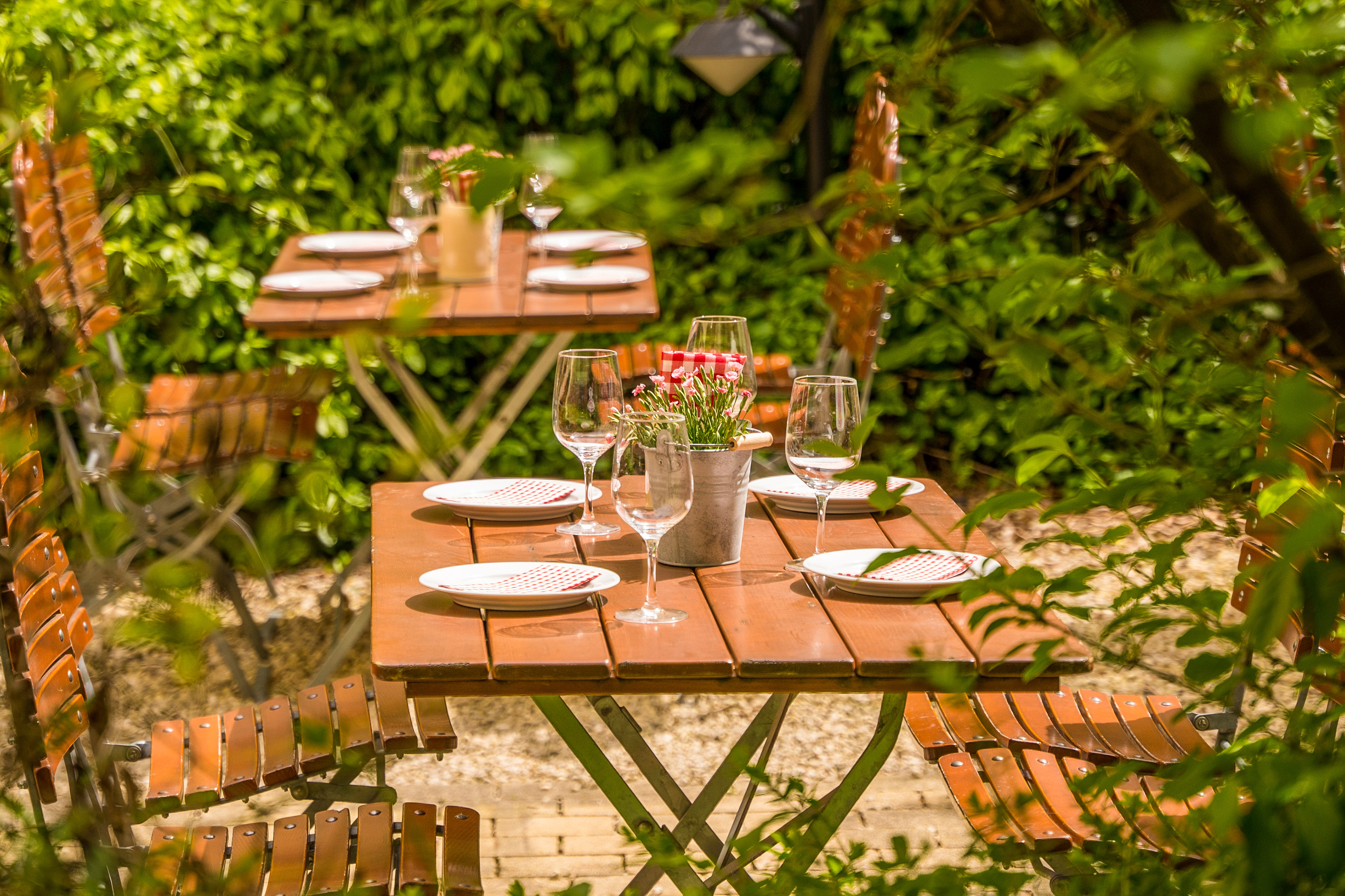 Al fresco dining tables and chairs in a green garden, set with glasses and plates.