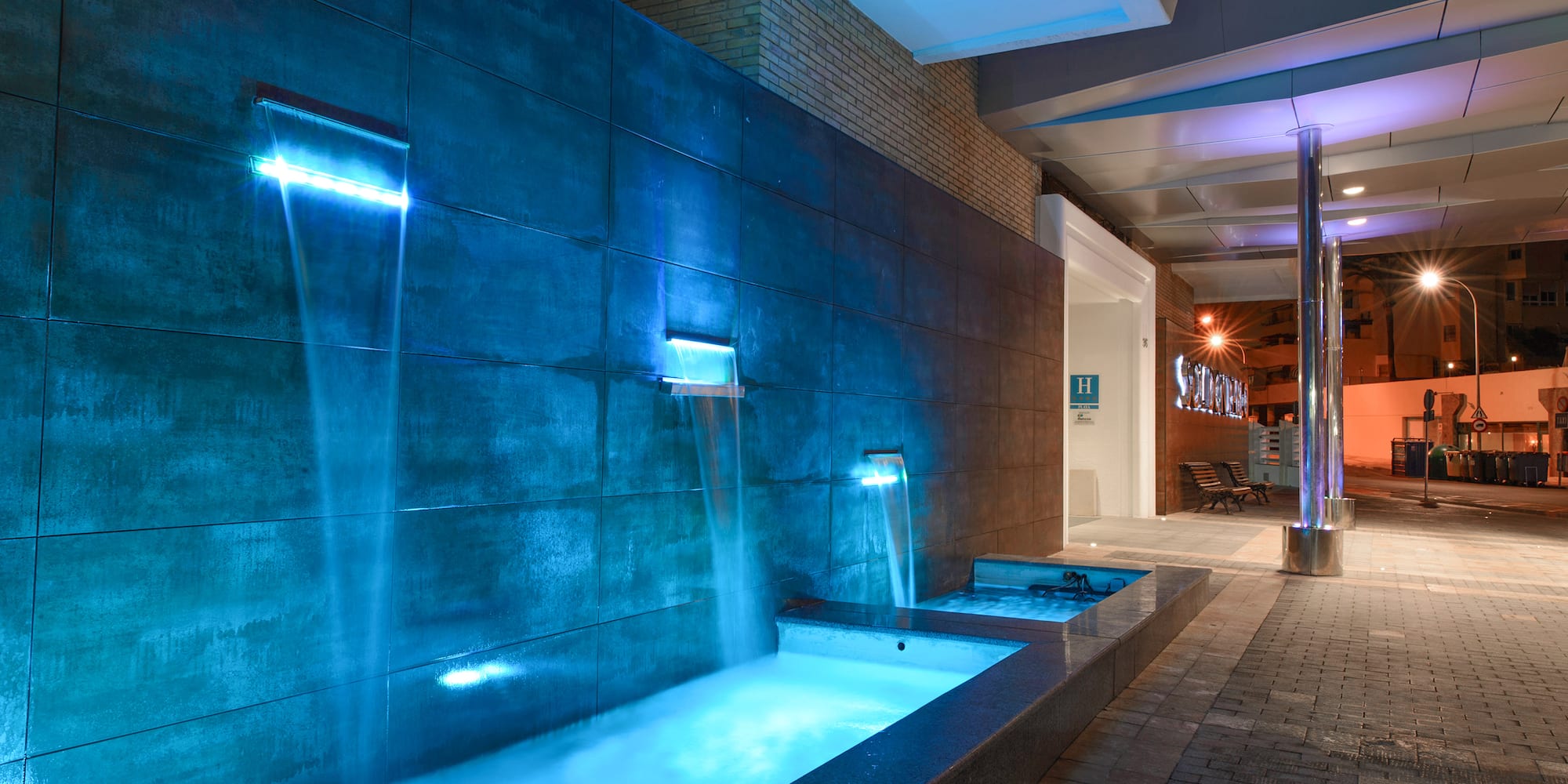 Blue illuminated waterfall at hotel entrance, modern tiled wall, night view.
