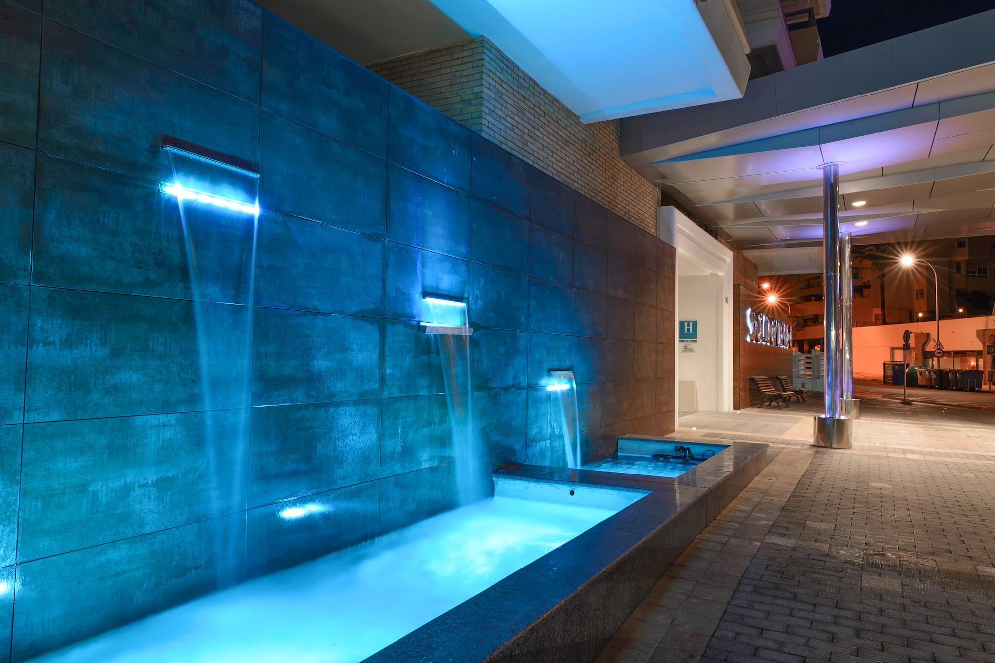 Blue illuminated waterfall at hotel entrance, modern tiled wall, night view.