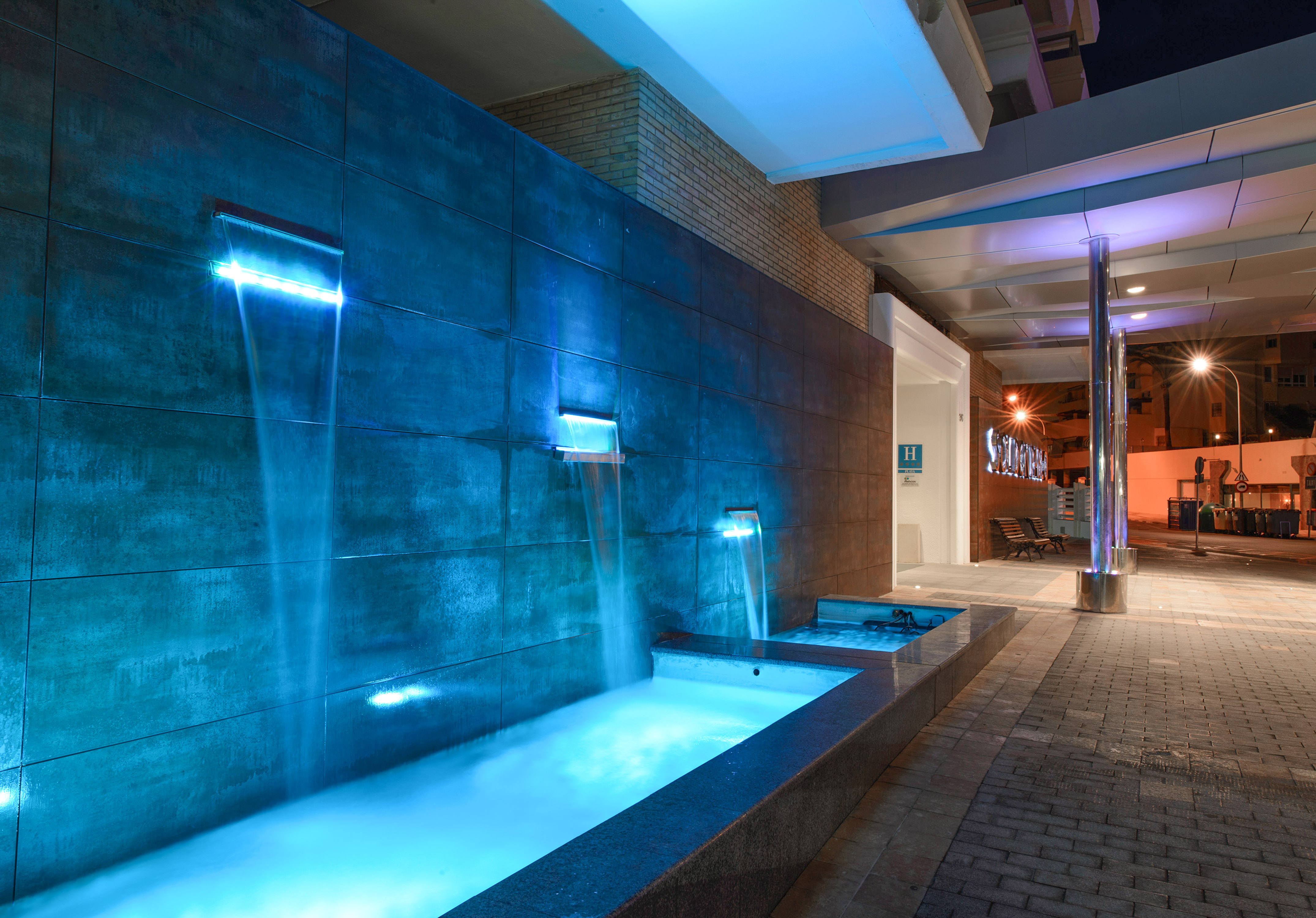 Blue illuminated waterfall at hotel entrance, modern tiled wall, night view.