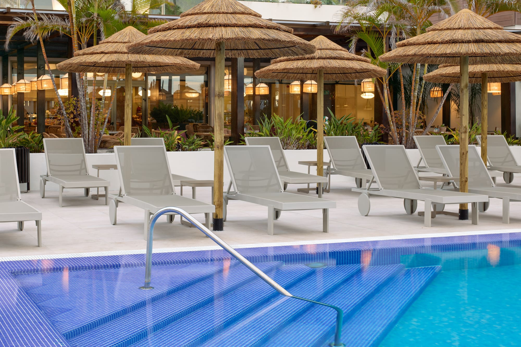 Resort swimming pool with blue tiles, lounge chairs, thatch umbrellas, palm trees.