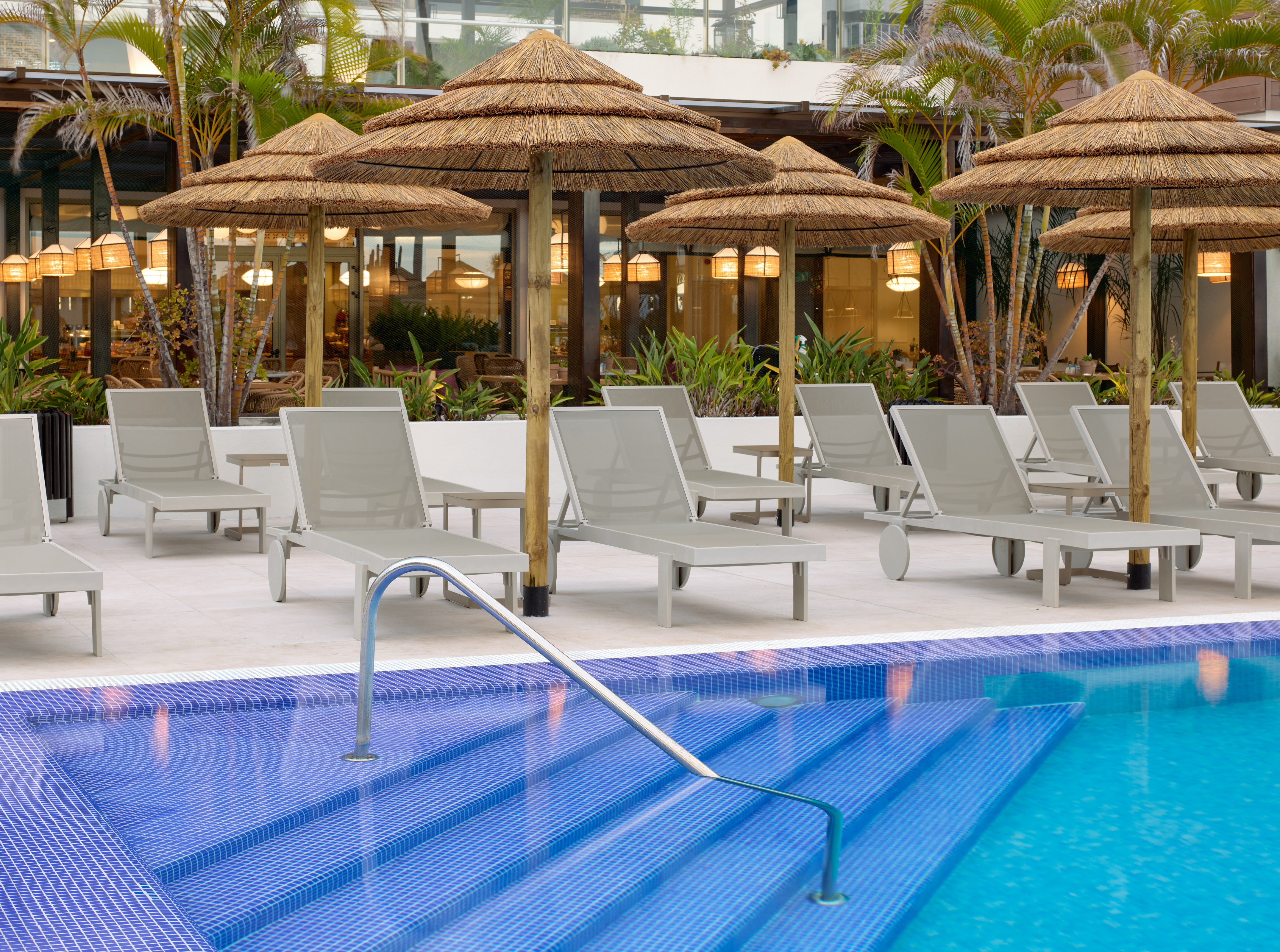 Resort swimming pool with blue tiles, lounge chairs, thatch umbrellas, palm trees.