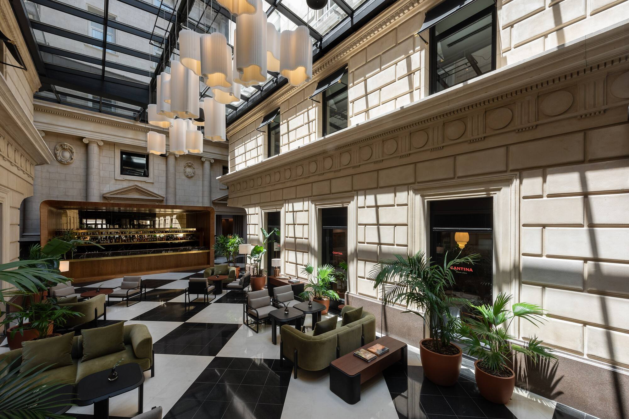 Hotel atrium lounge with glass roof, chandeliers, checkerboard floor, and bar.