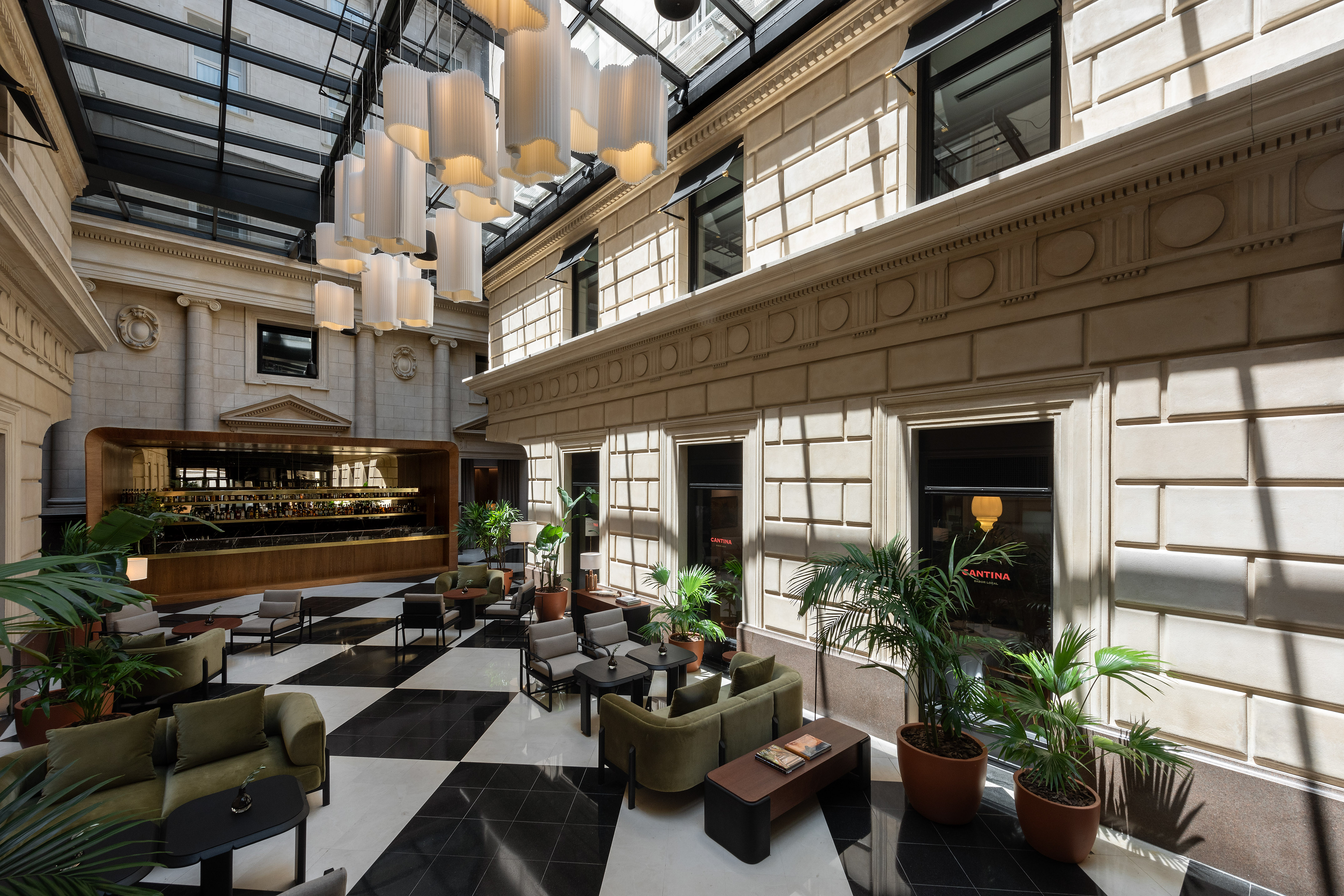 Hotel atrium lounge with glass roof, chandeliers, checkerboard floor, and bar.