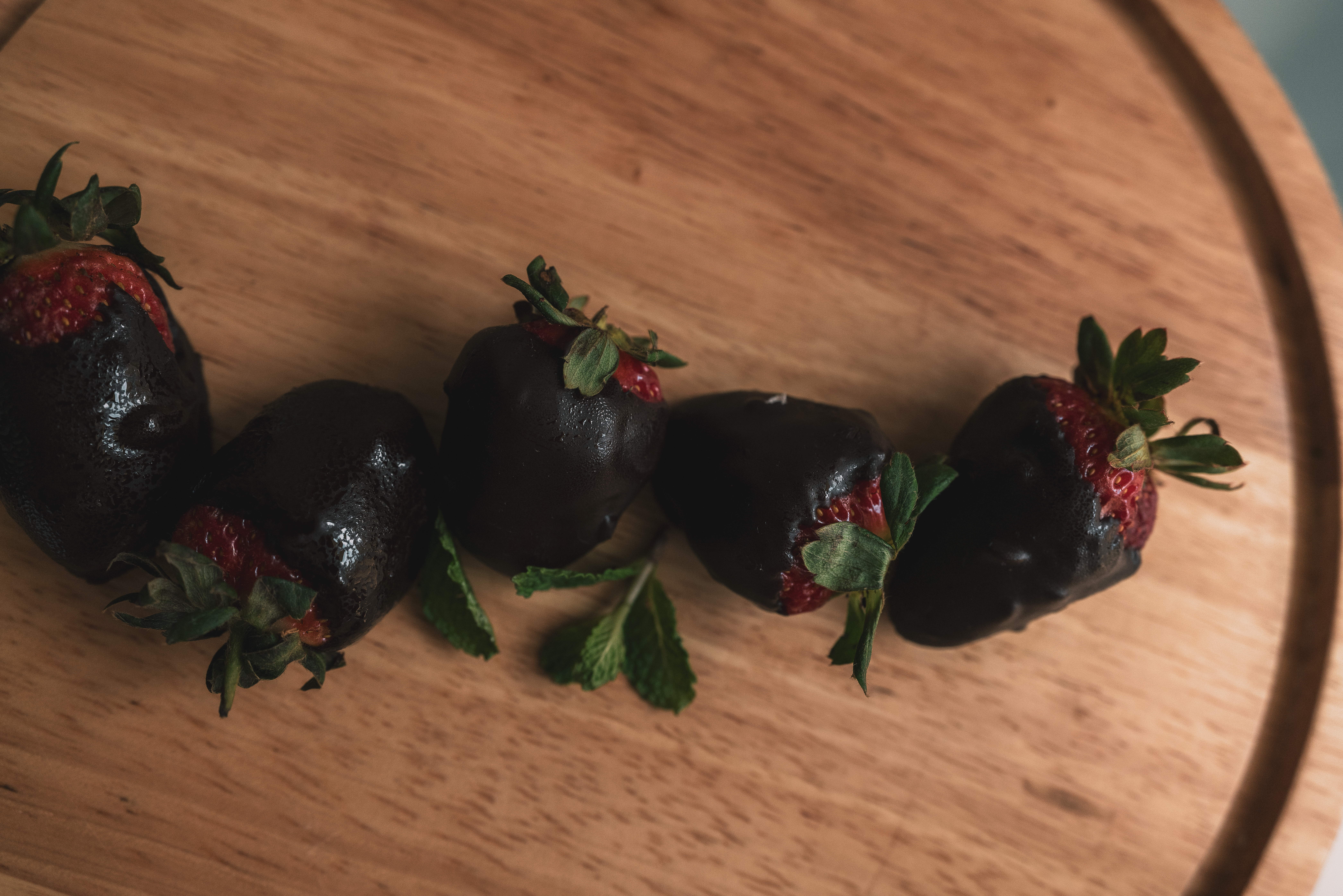Four dark chocolate-covered strawberries with fresh mint on a wooden board.